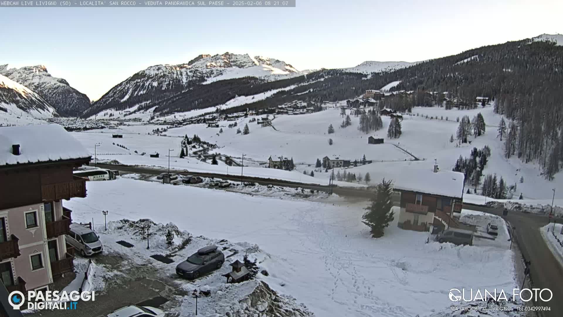 A snow-covered mountain valley village scene on a clear, bright day, with buildings and parked cars visible in the foreground.