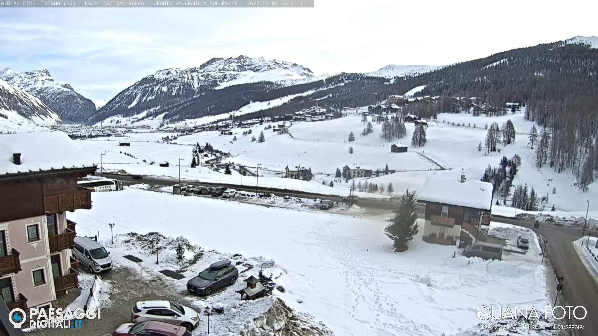 A snow-covered mountain village scene, viewed from a slightly elevated position, shows several buildings and cars parked in the snow under a cloudy sky.