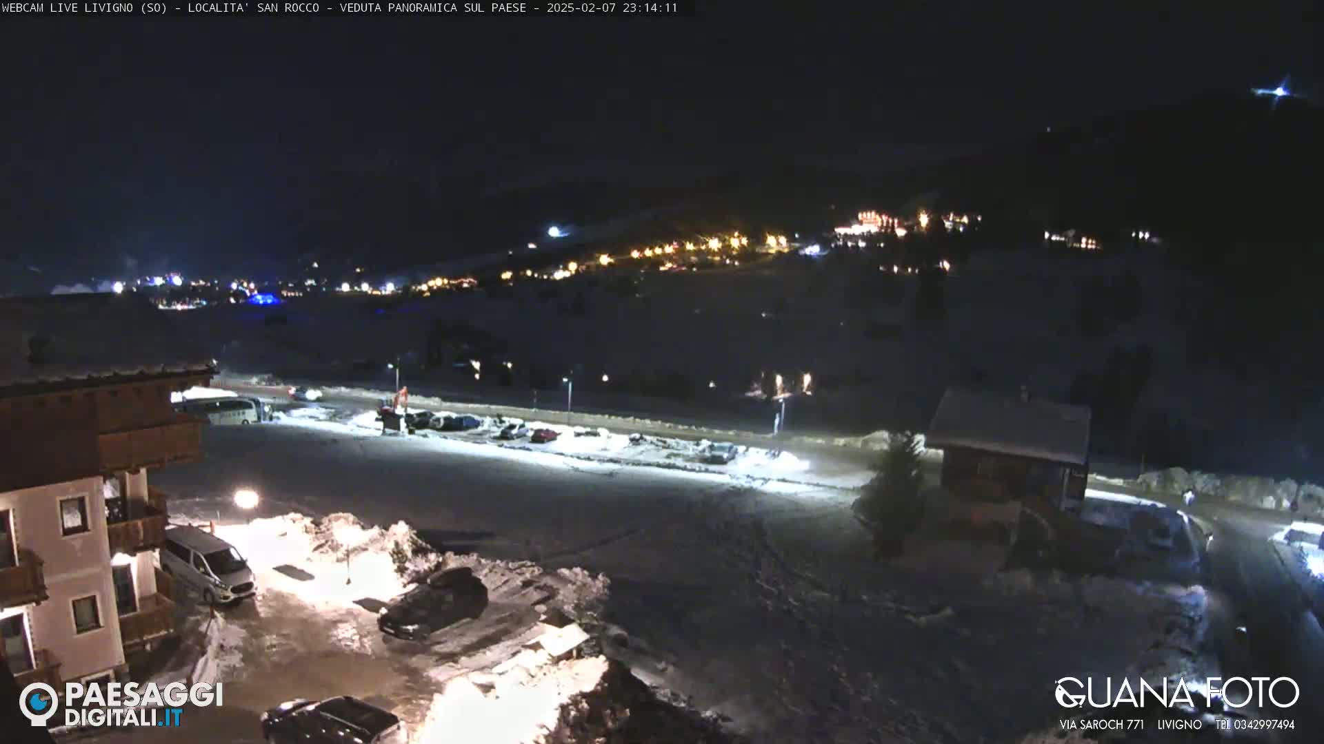 A nighttime, snow-covered parking lot and surrounding buildings are illuminated by artificial lights, with a small village visible in the distance under a dark sky.