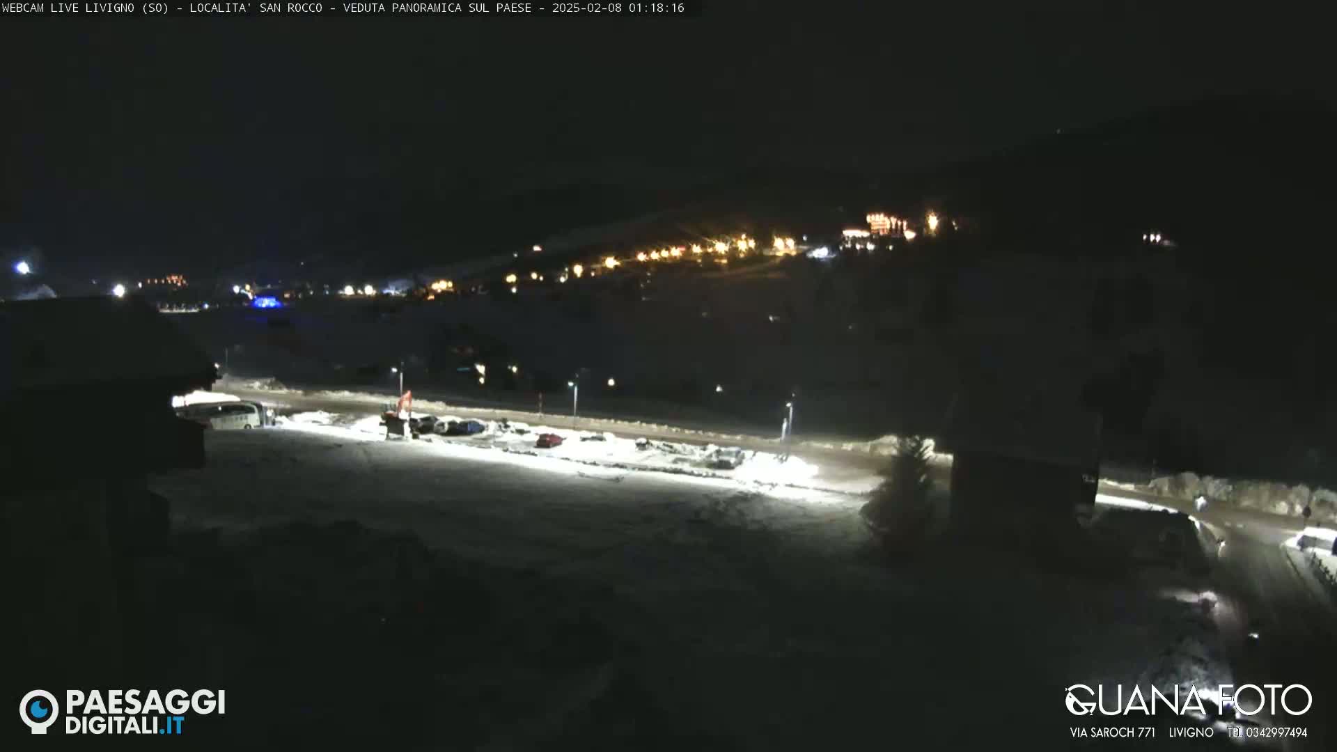 A snow-covered parking lot at night, illuminated by artificial lights, with a village visible in the background under a dark sky.