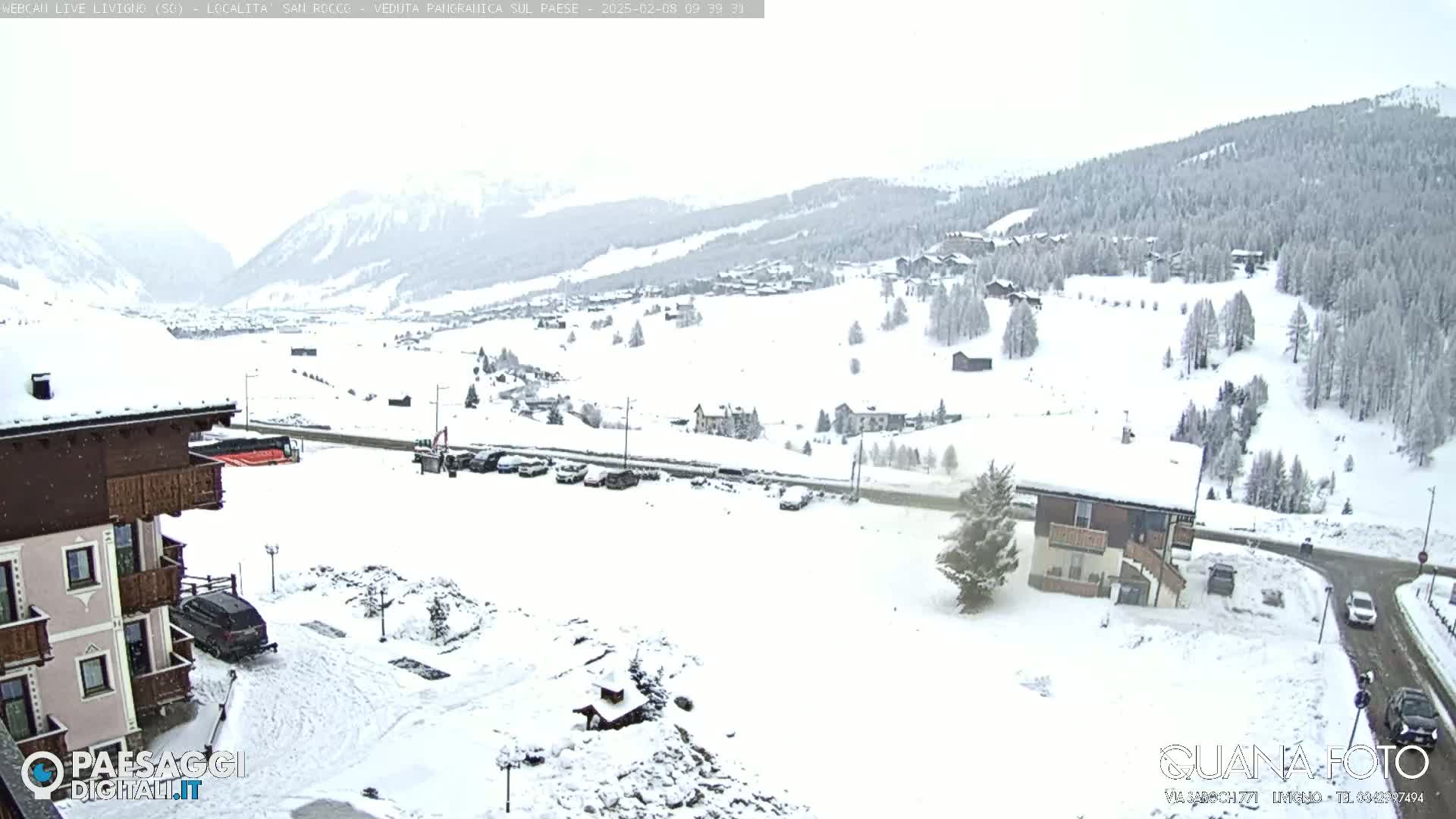 A snow-covered village nestled in a valley between snow-dusted mountains, with a building in the foreground and a few cars visible in a parking lot.