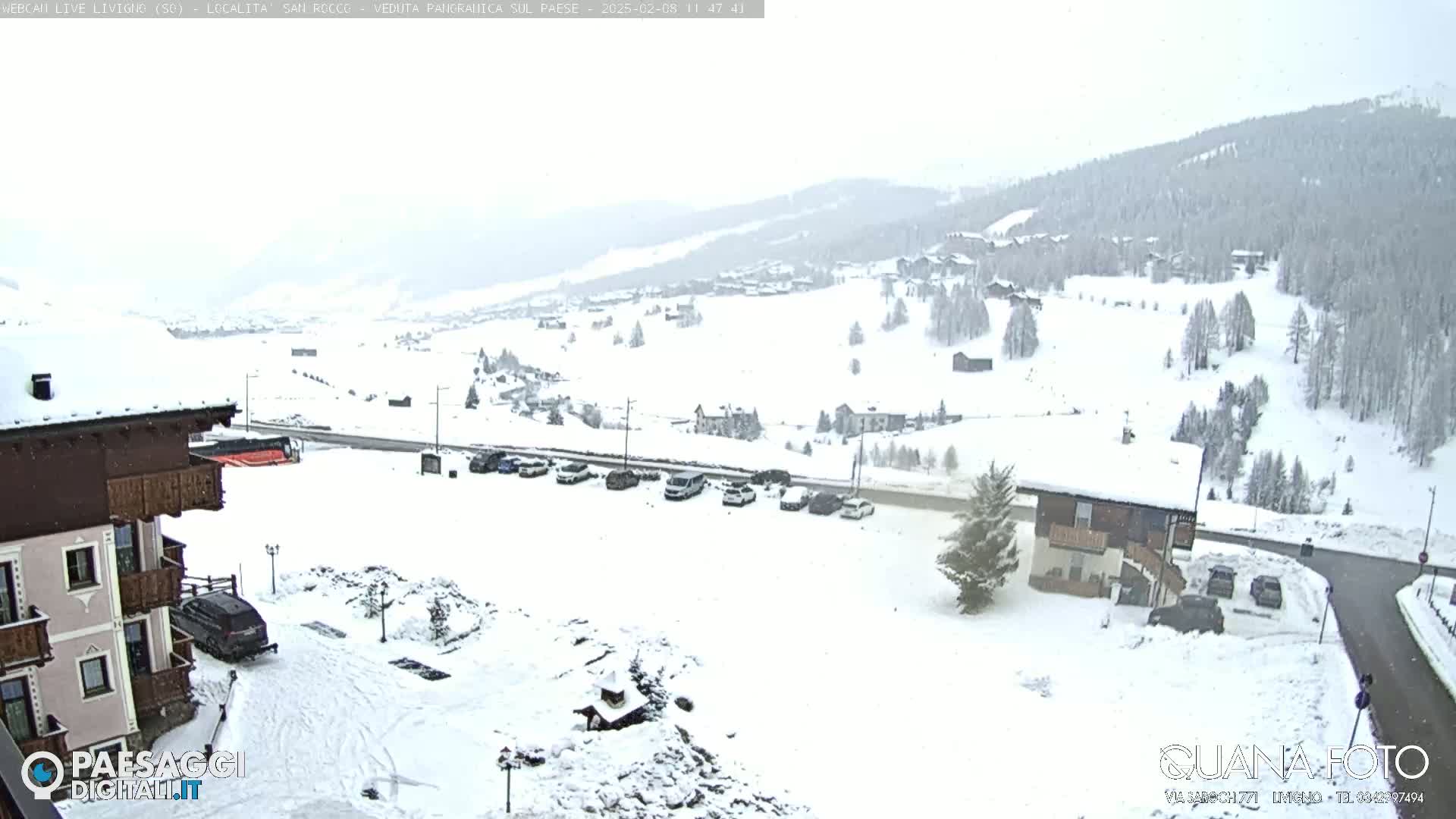 A snow-covered mountain village scene is visible from a high vantage point, showing several buildings, parked cars, and a road winding through the landscape under a lightly snowing sky.