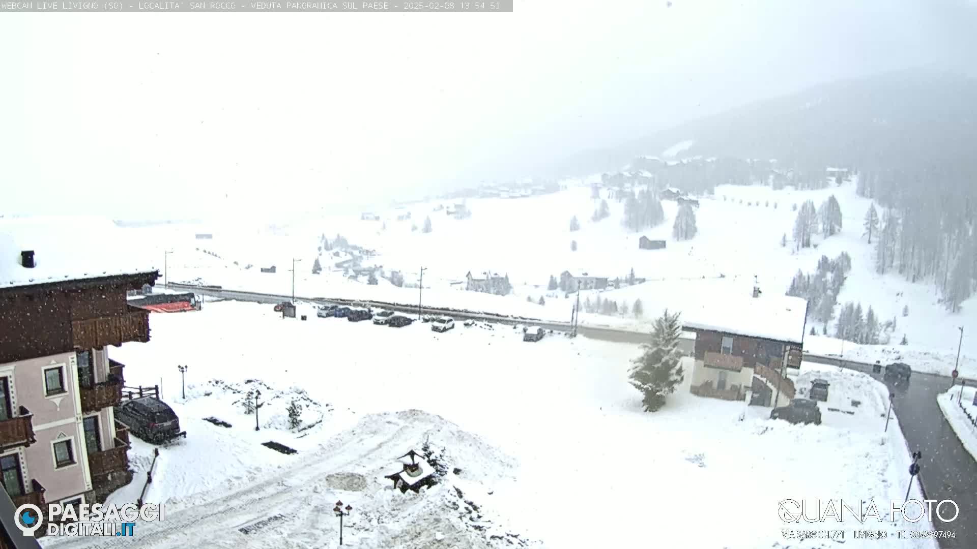 A snow-covered mountain village is seen during a light snowfall, with cars parked in a lot near a building.