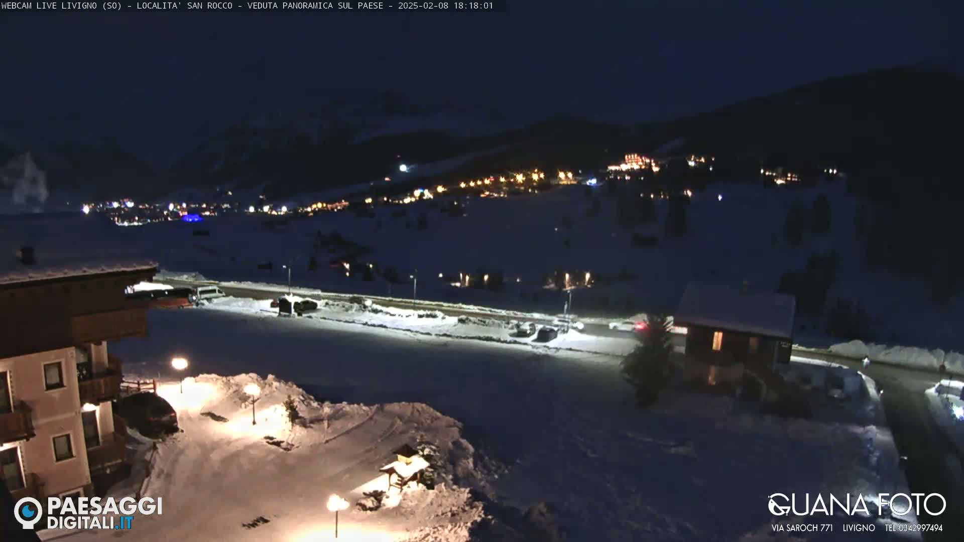 A nighttime, snowy scene shows a village in the distance illuminated by lights, with a snow-covered road and buildings in the foreground under a dark sky.