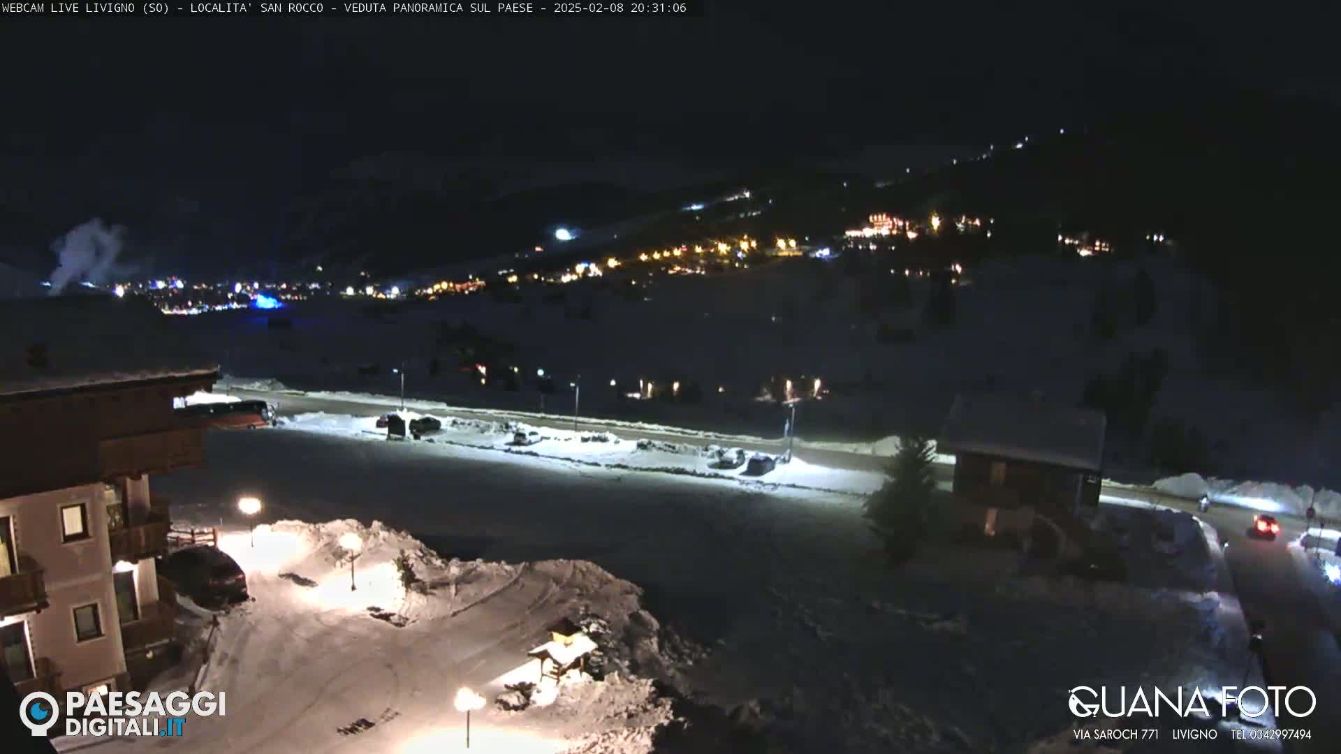 A nighttime, snowy scene shows a town illuminated in the distance with a few cars and buildings visible in the foreground under dark, clear skies.