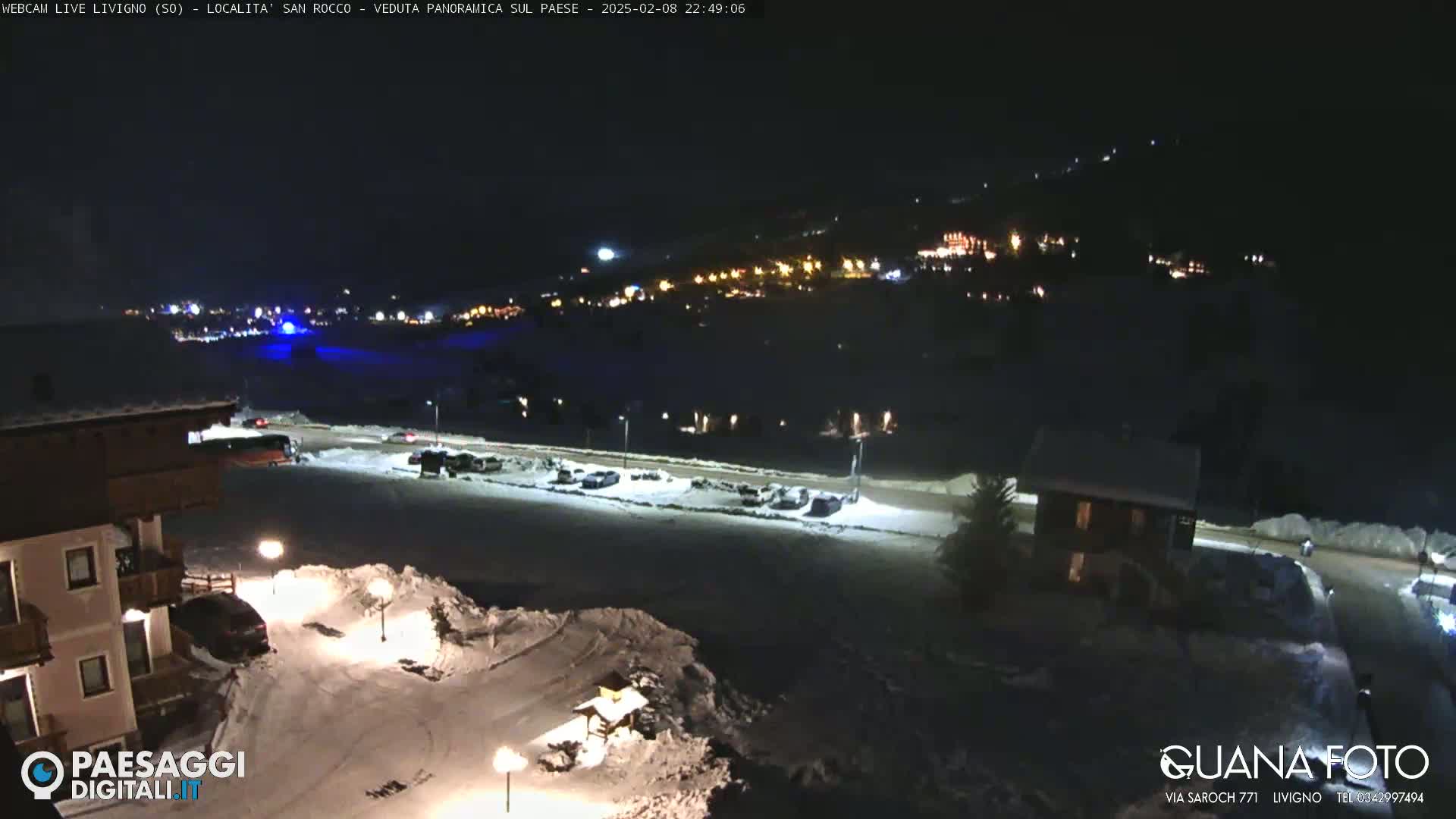 A nighttime, snow-covered scene shows a village illuminated in the distance, with a parking lot and buildings in the foreground under artificial lights.