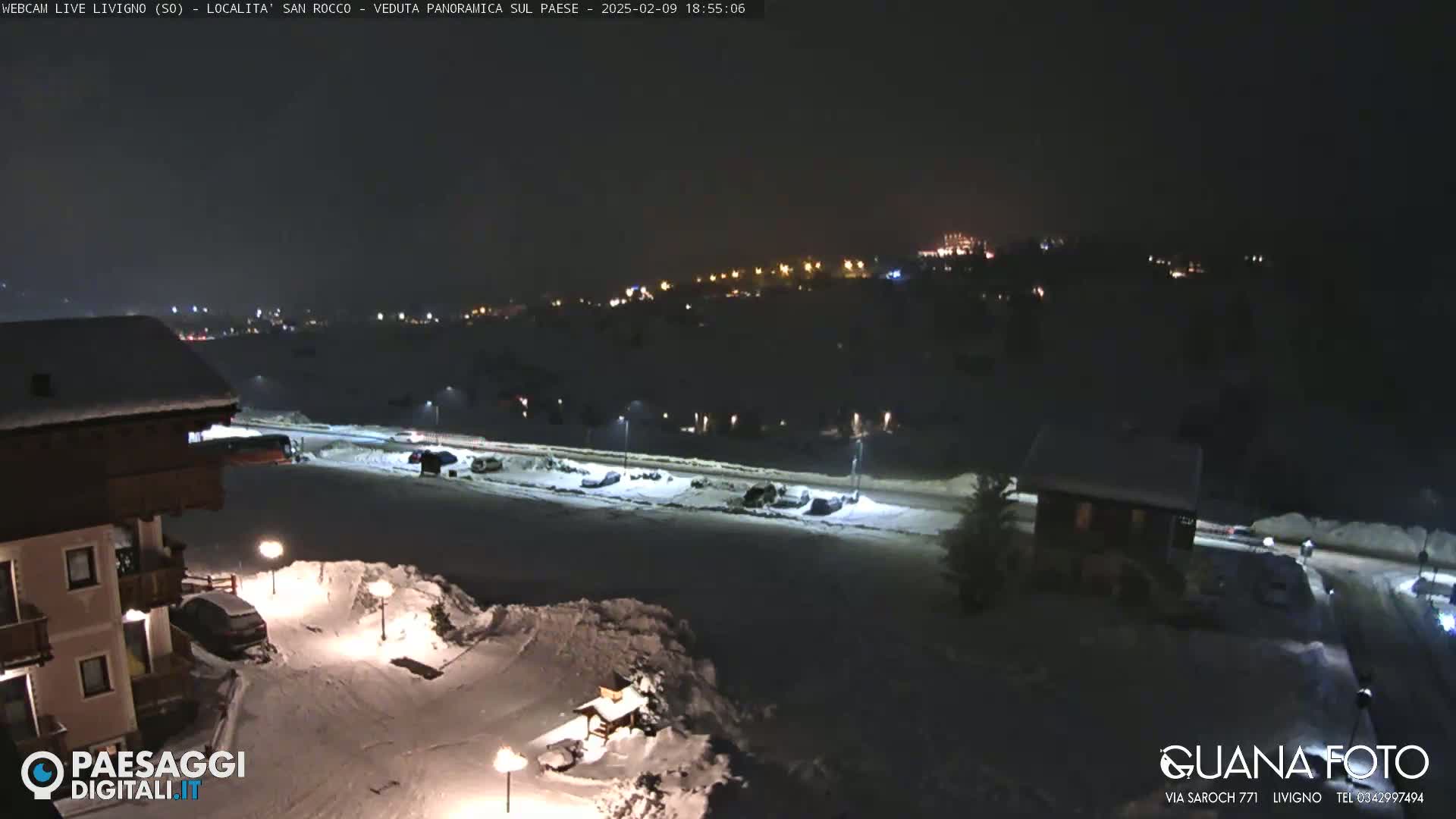 A snow-covered nighttime scene shows a parking lot with cars, buildings, and distant lights on a hillside under a dark sky.