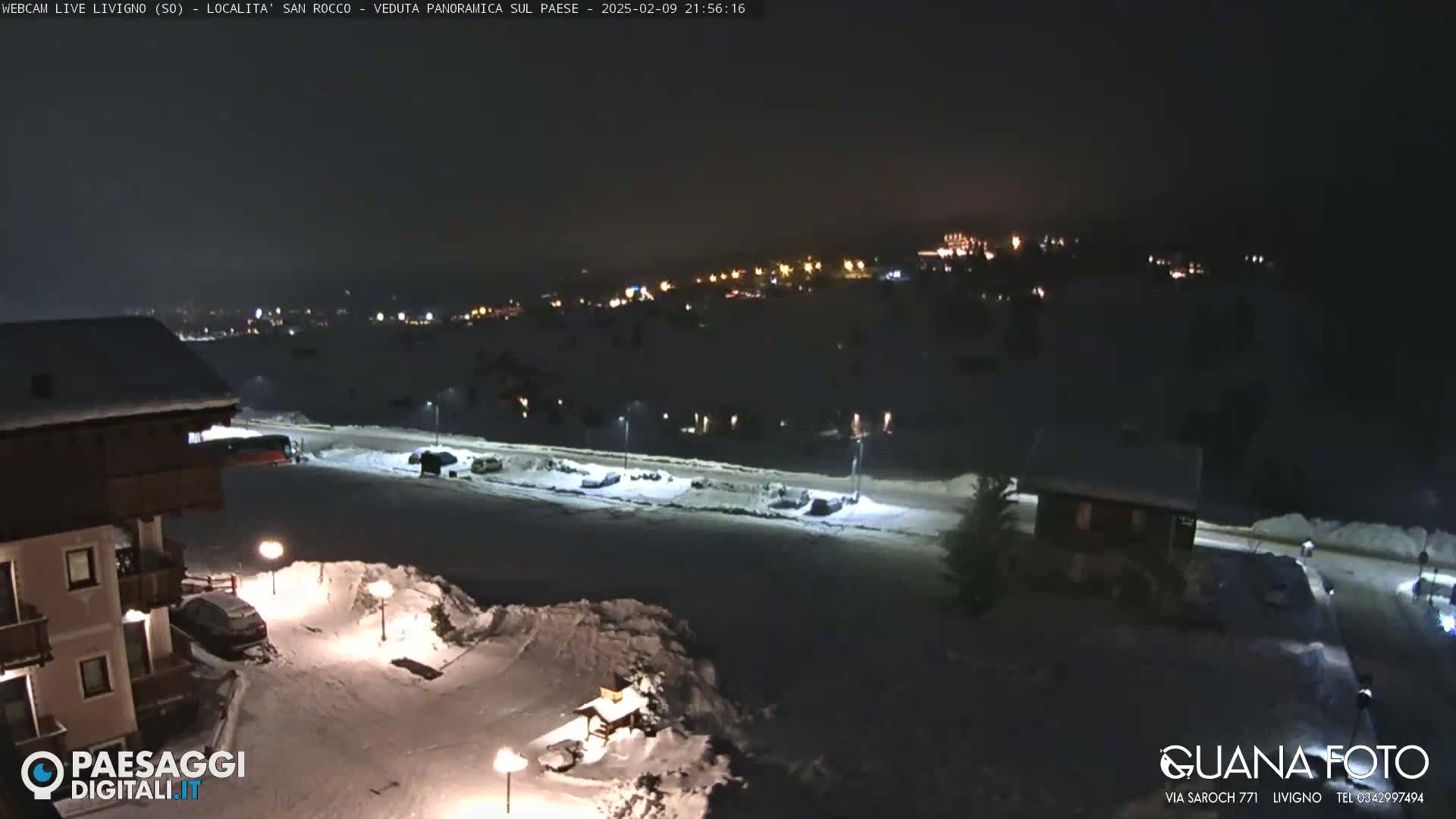 A snow-covered nighttime scene shows a village with scattered lights, a building in the foreground, and parked cars along a road.