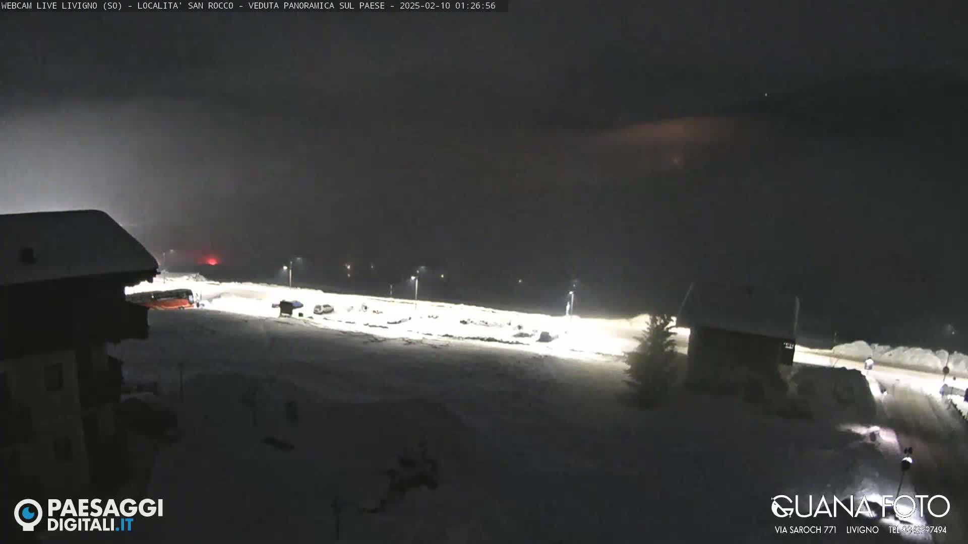 A snow-covered parking lot and buildings are illuminated at night under a dark, possibly cloudy sky.