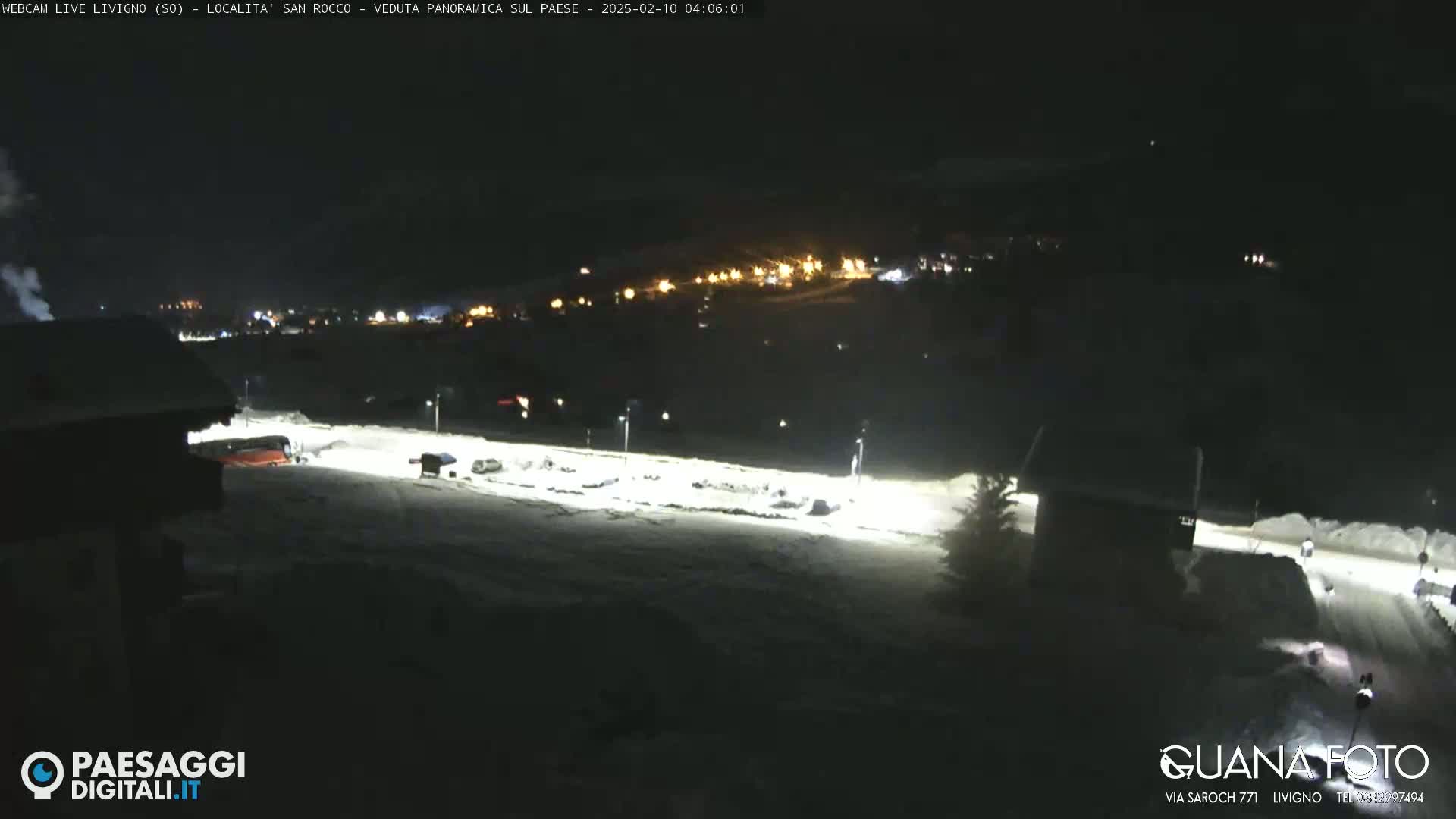 A snow-covered nighttime scene shows a village's lights in the distance, with a few vehicles parked in a snow-covered lot in the foreground.