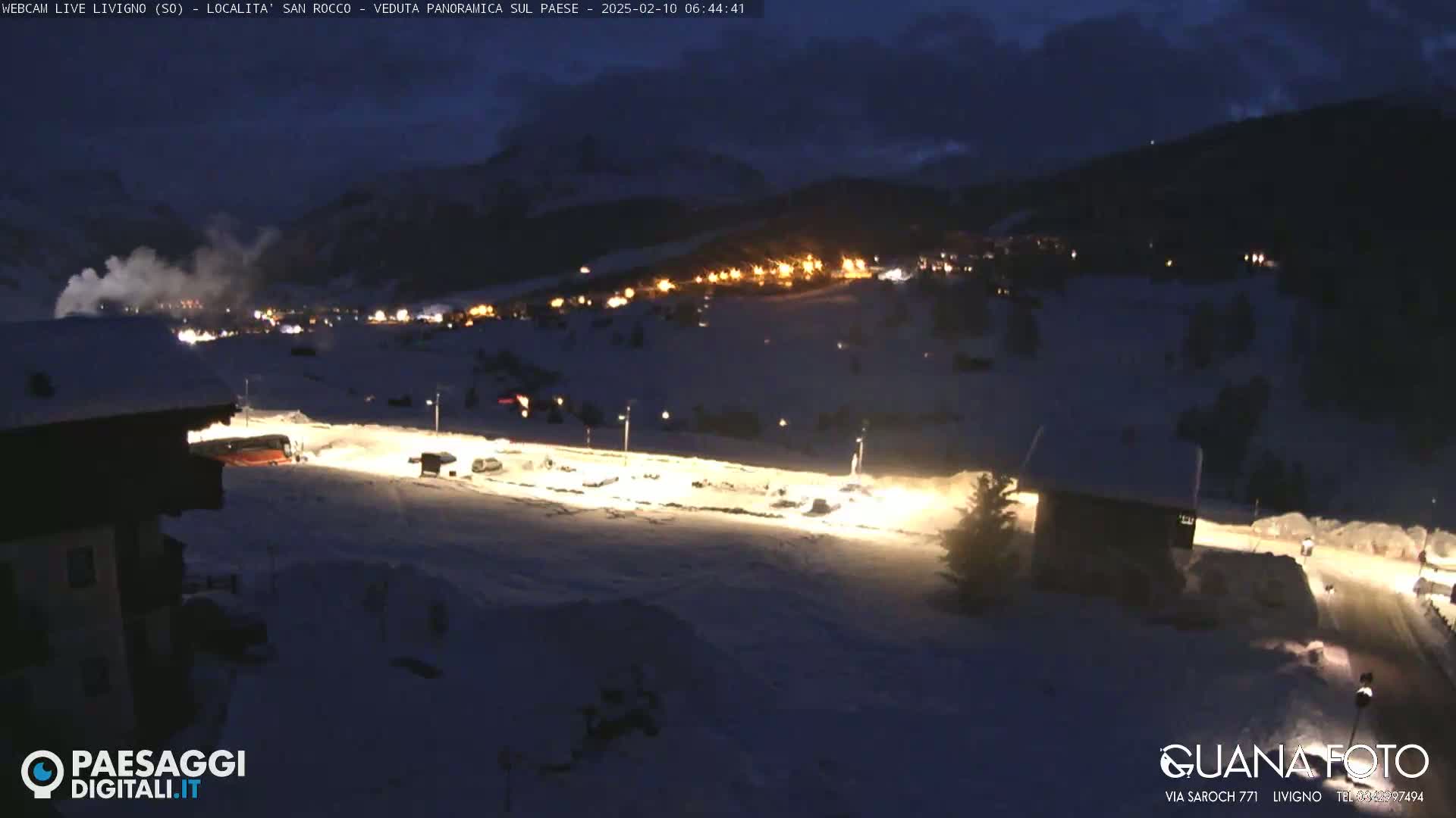 A snow-covered landscape at night, illuminated by the lights of a village nestled in the mountains under a dark, cloudy sky.