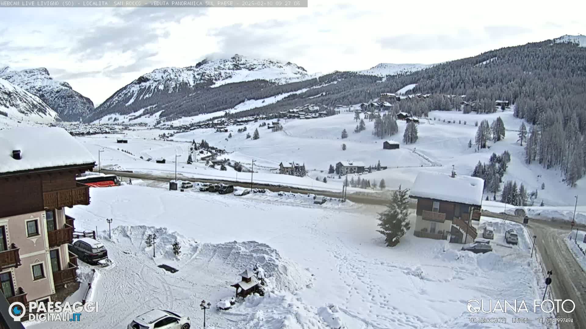 A snow-covered mountain village is viewed from an elevated position on a cloudy day, with cars parked in a snowy lot in the foreground.