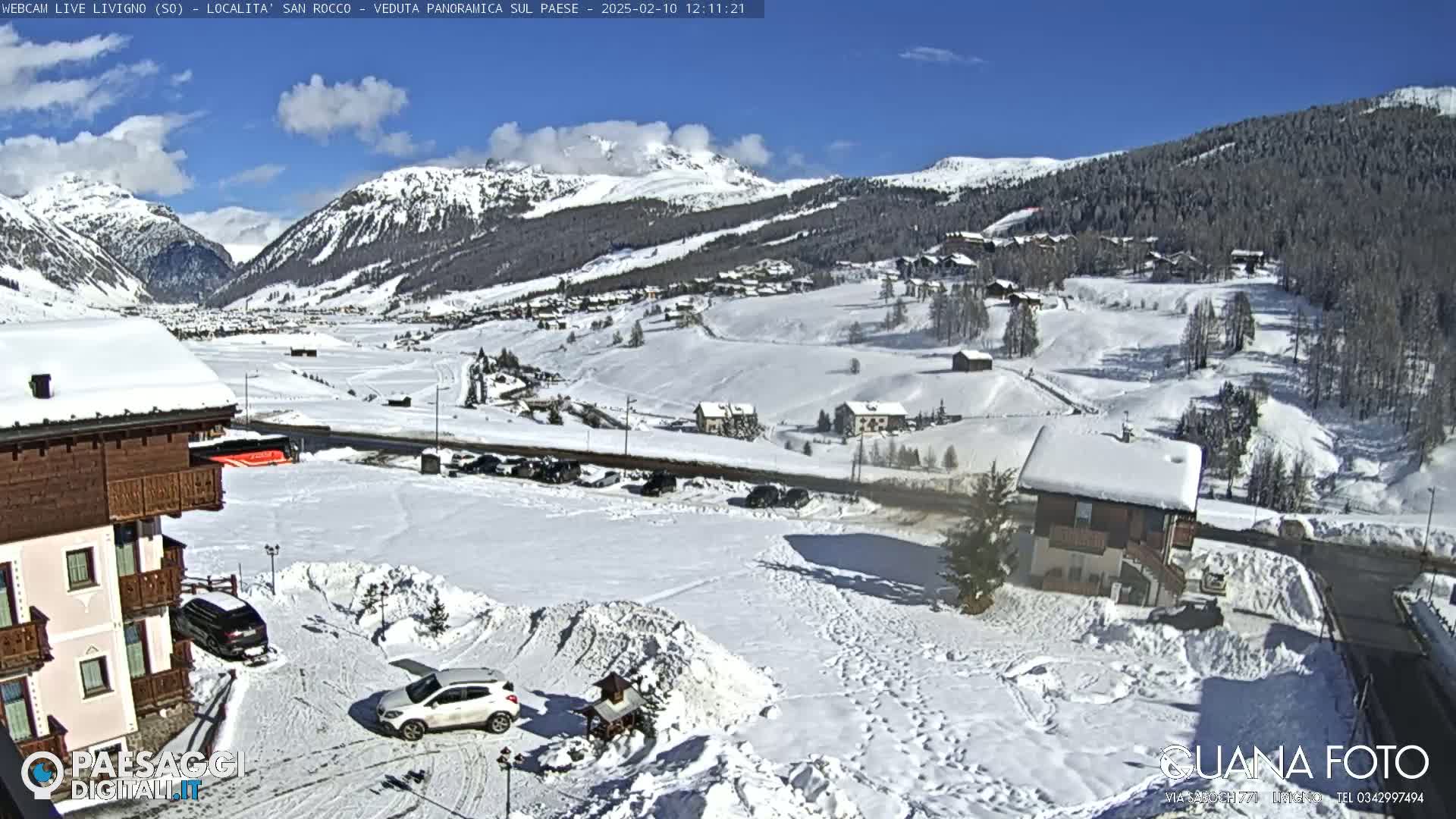 A snow-covered village nestled in a valley between snow-capped mountains under a mostly sunny sky.