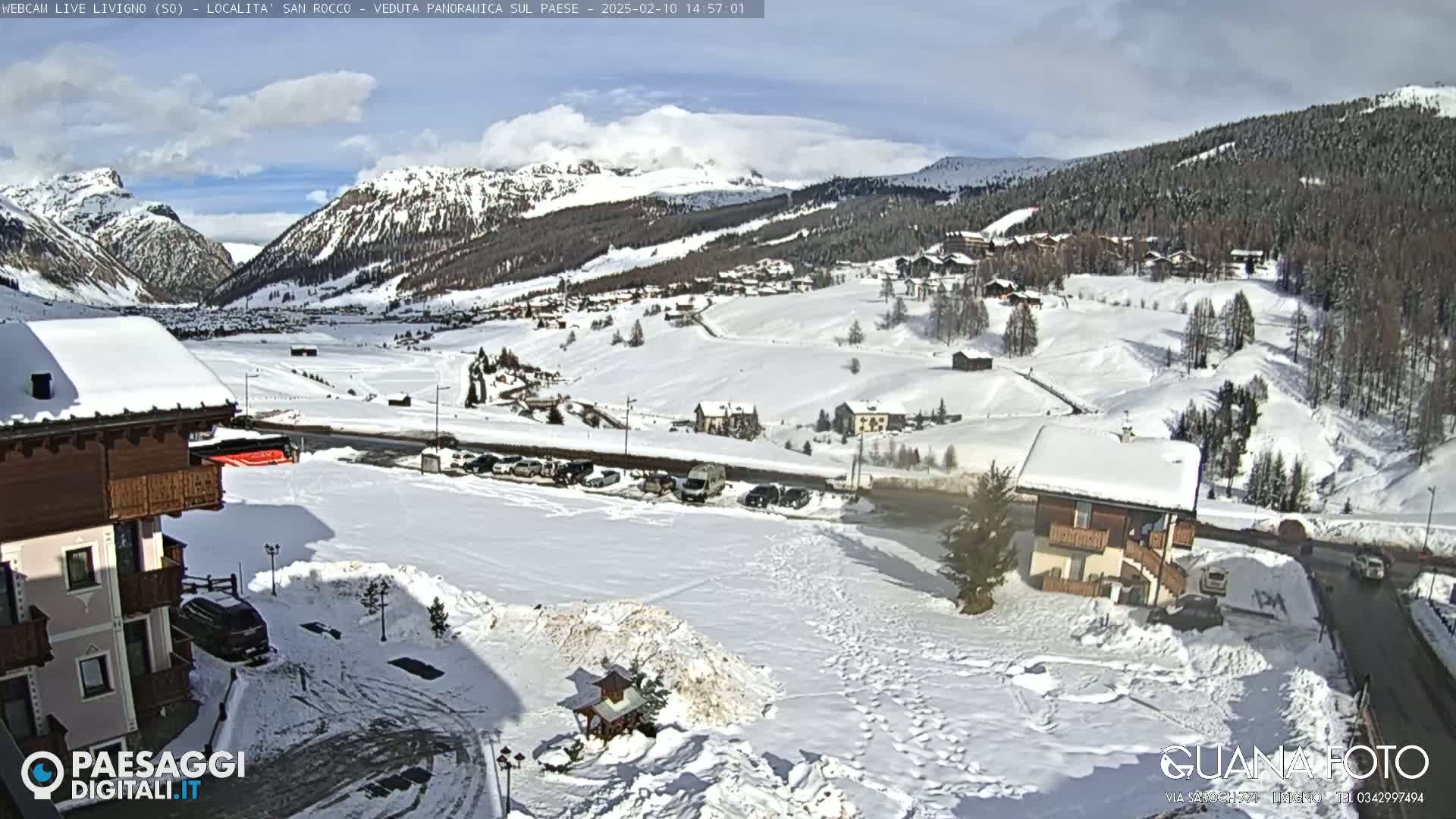 A snow-covered mountain village is viewed from a hilltop, under a mostly sunny sky with some clouds.