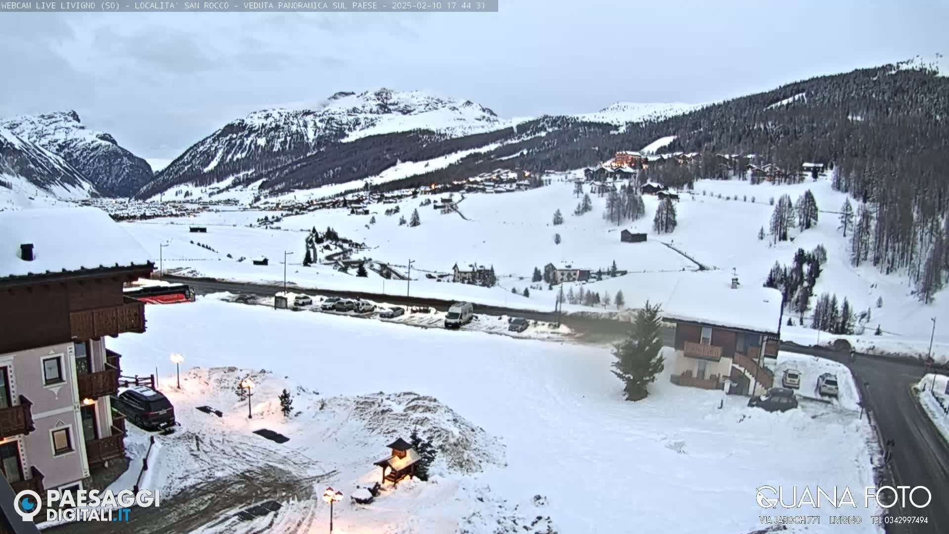 A snow-covered mountain village is viewed from a hillside, with overcast skies and a building in the foreground.