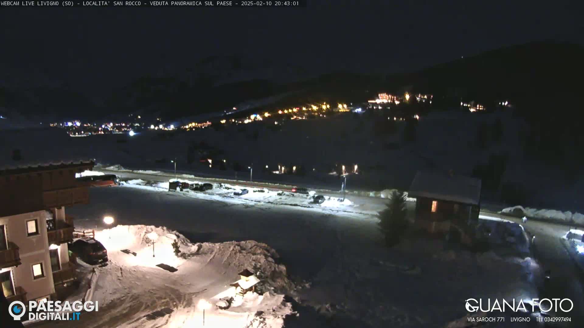 A nighttime, snow-covered scene shows a village with lights in the distance, a parking lot with cars, and a building in the foreground under a dark sky.