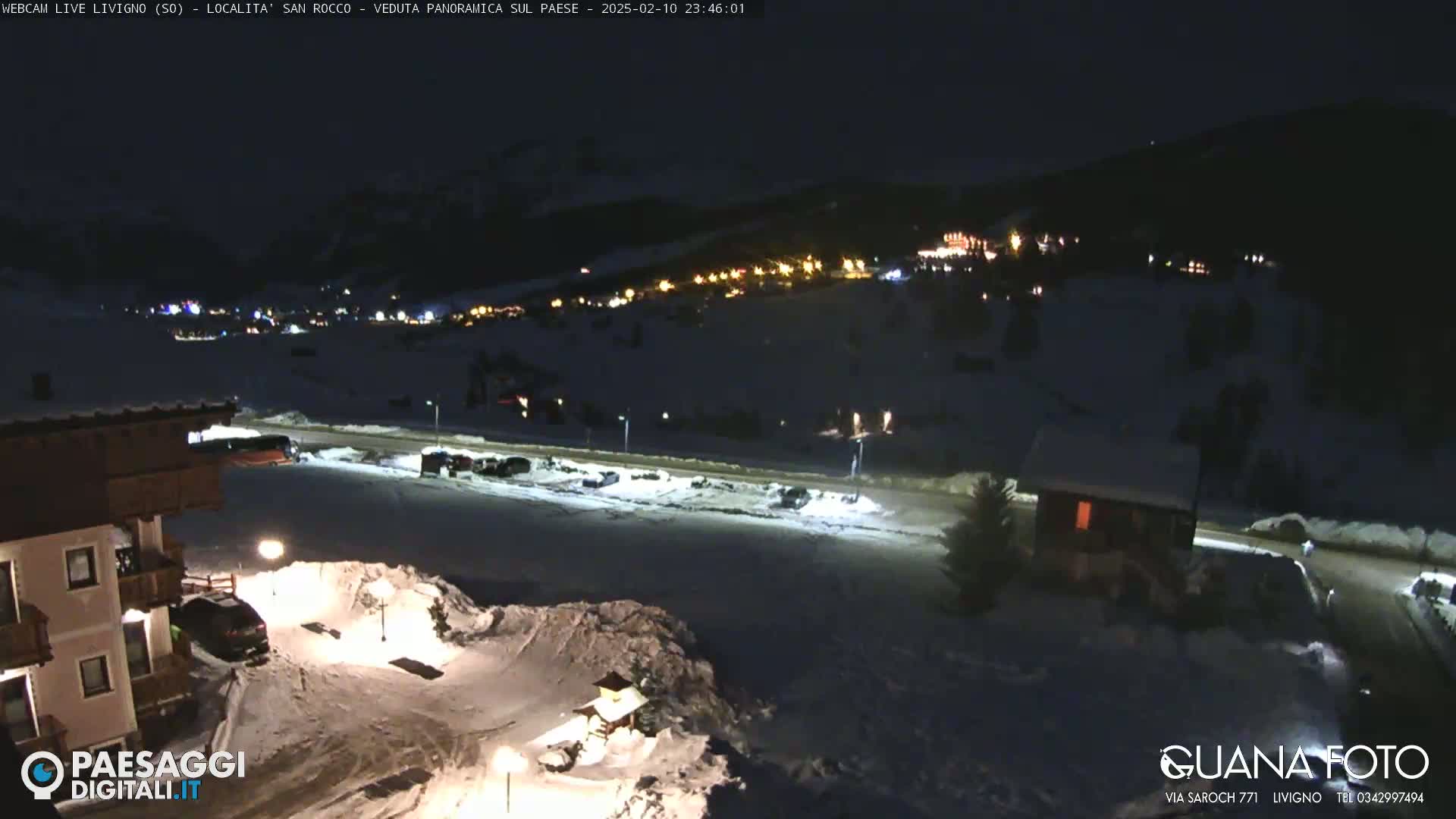A nighttime, snow-covered village scene is illuminated by streetlights and building lights, with a partially visible building in the foreground.