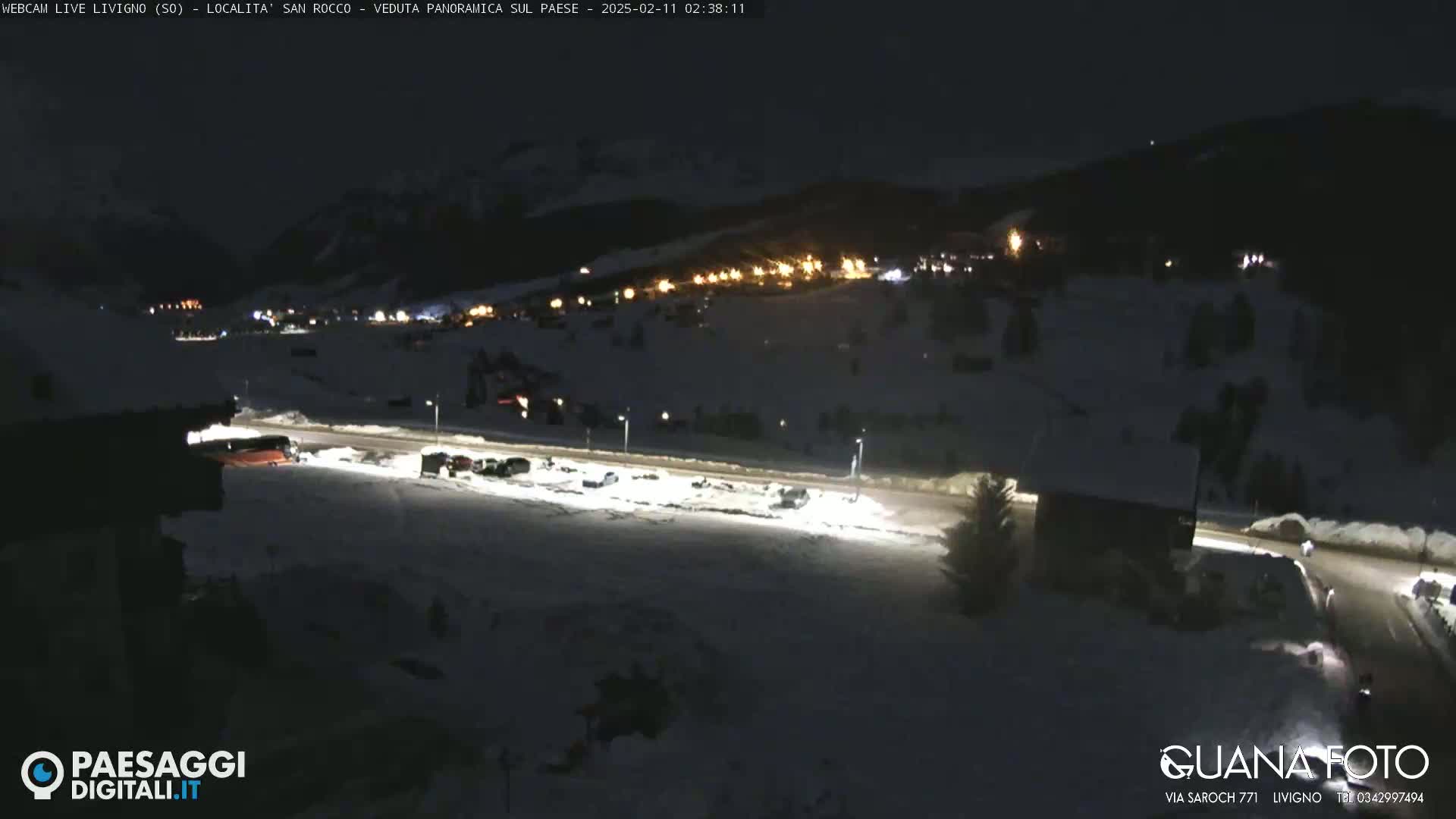 A nighttime, snow-covered parking lot and road are illuminated by streetlights, with a village and distant mountains visible in the background under a dark sky.