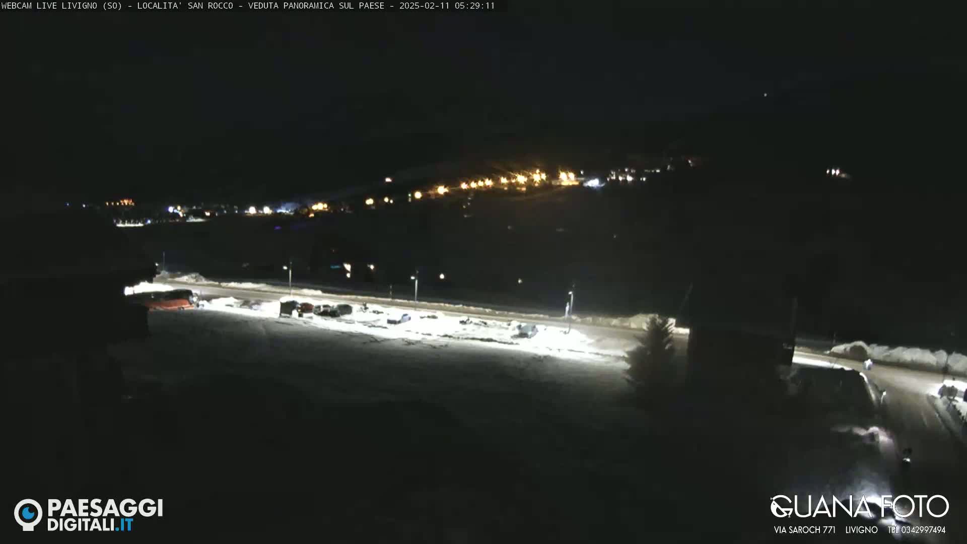 A nighttime, snowy scene shows a mostly empty parking lot with a few vehicles, illuminated by bright lights, and a distant town with lights visible in the background.