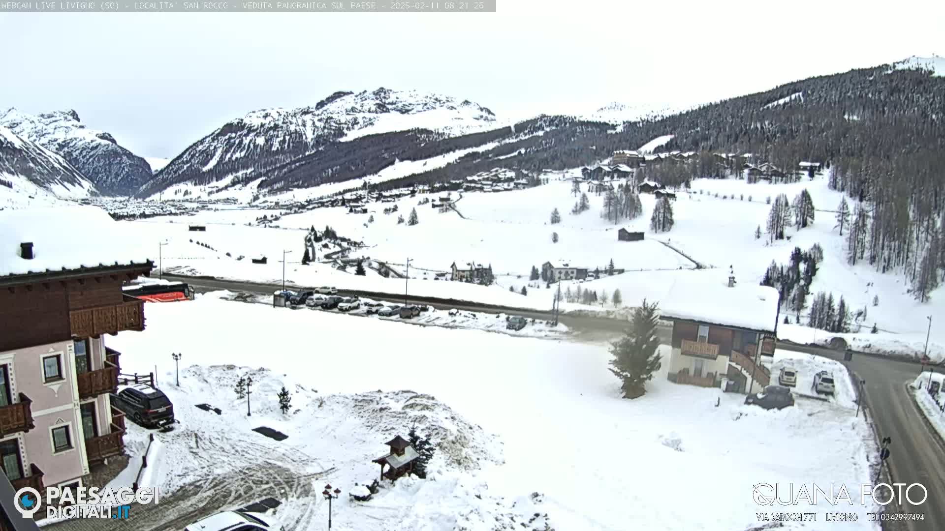 A snow-covered mountain valley village is seen on an overcast day, with a building in the foreground and cars parked in a snow-covered lot.