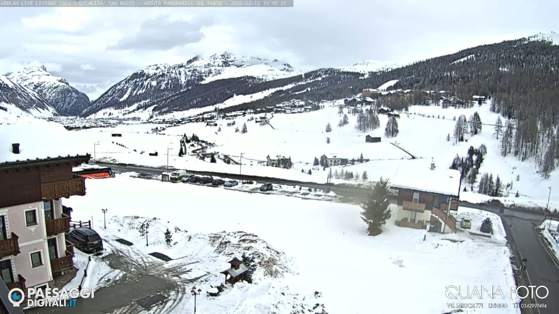 A snow-covered mountain village is viewed from an elevated position on an overcast day, with a few buildings and cars visible in the foreground.