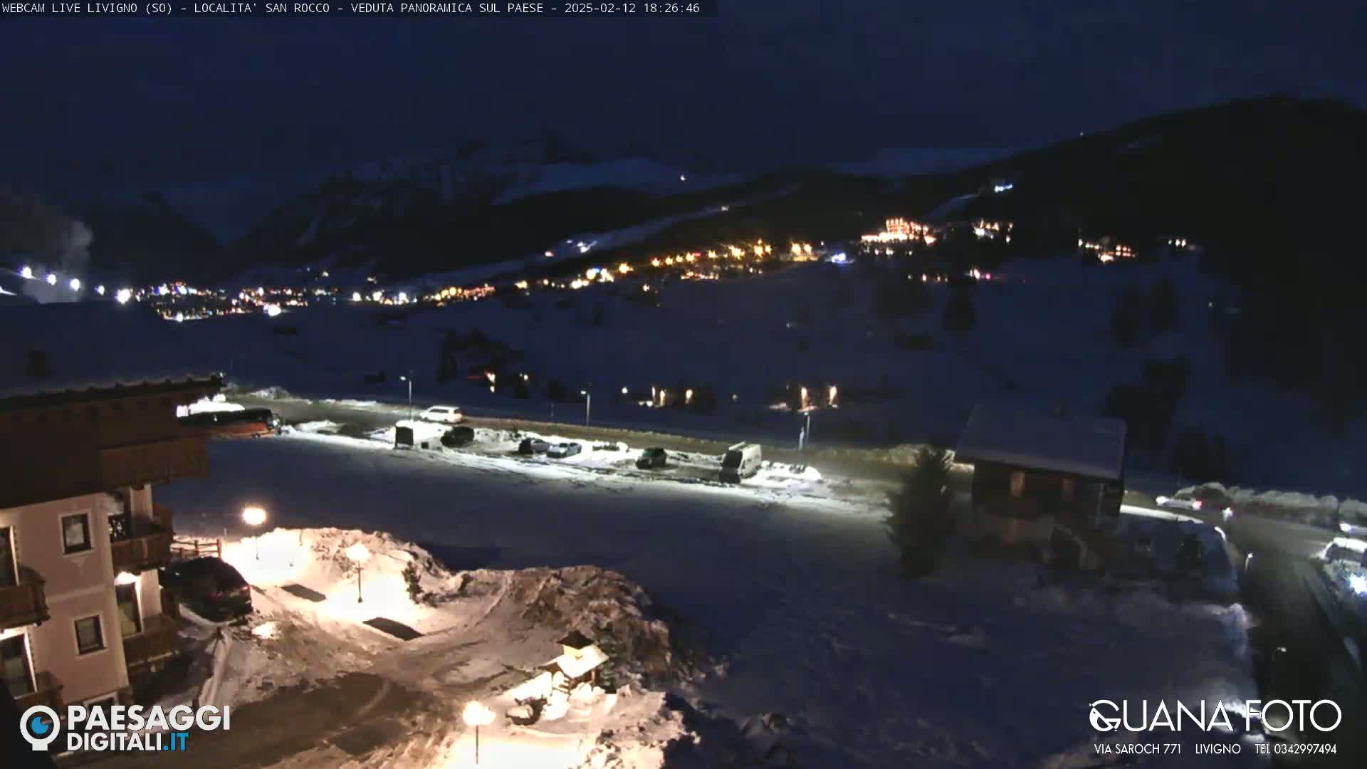 A nighttime, snowy scene shows a small village nestled in a valley, illuminated by lights, with a parking lot and buildings in the foreground.