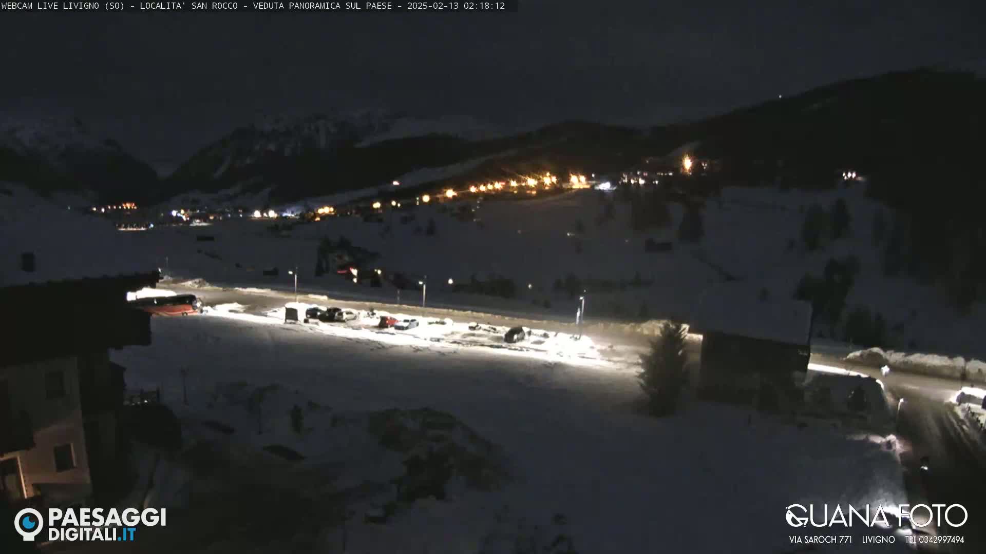 A snow-covered parking lot with several cars and a bus is illuminated at night, overlooking a town nestled in a dark, snow-covered mountain valley.