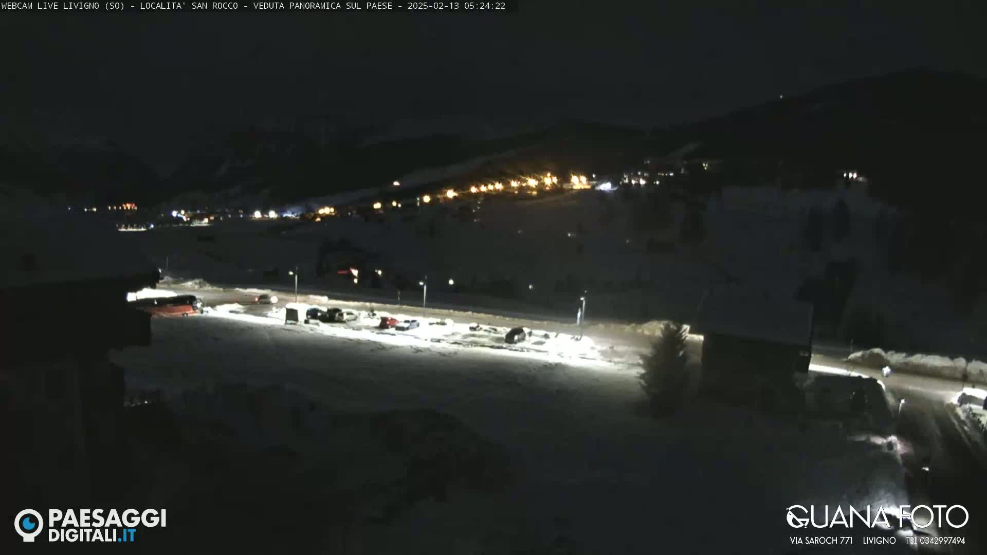 A nighttime, snow-covered parking lot with several vehicles and a small building, overlooked by a distant village with illuminated buildings under a dark sky.