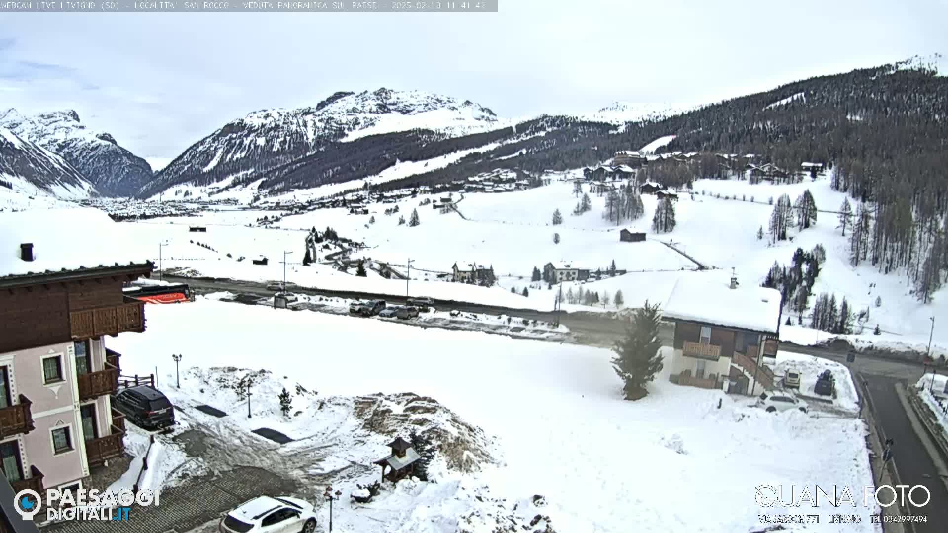 A snow-covered valley with mountains in the background, viewed from a slightly elevated position near some buildings and cars.