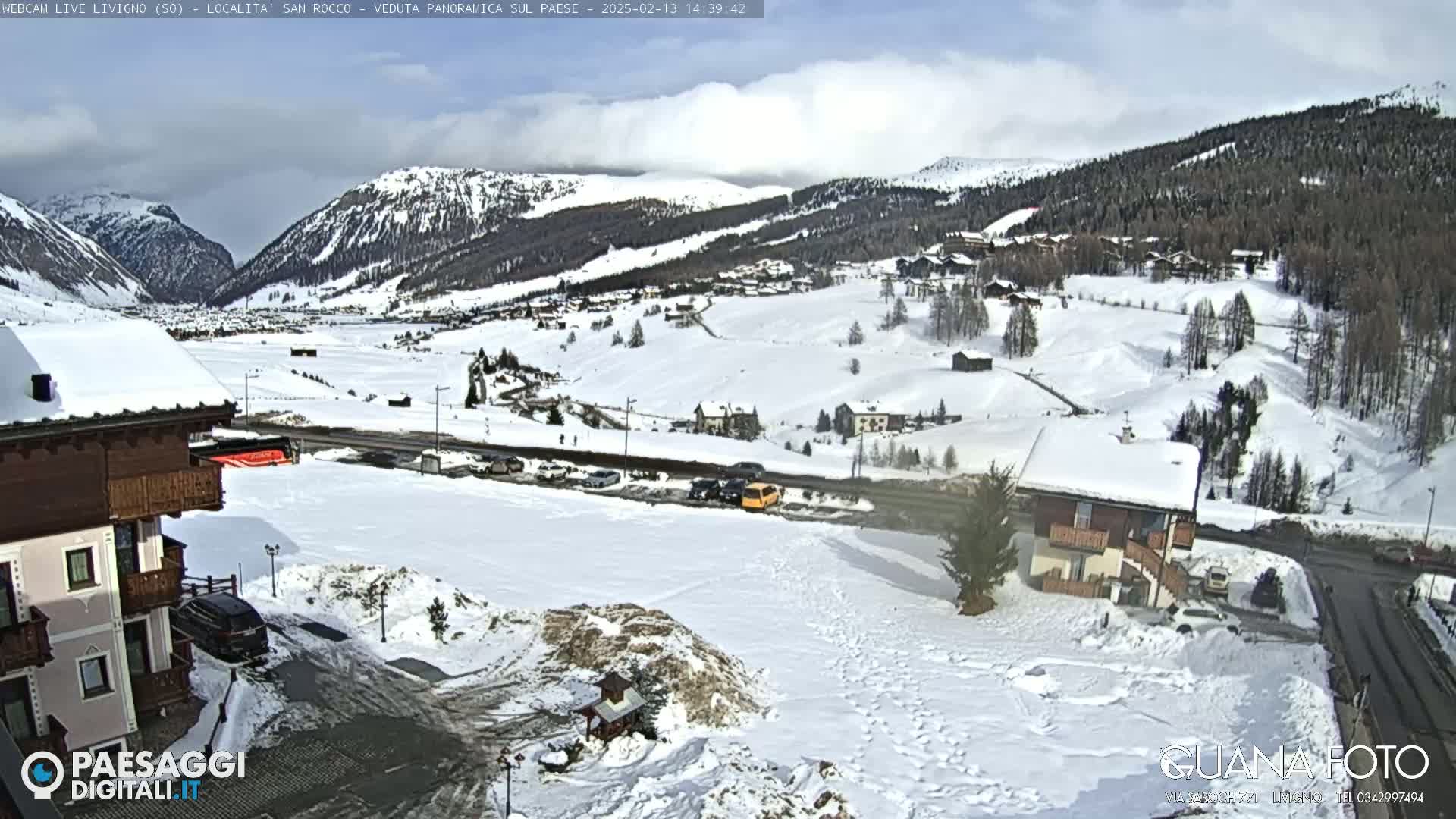 A snow-covered mountain village is viewed from a hilltop on a sunny day, with several buildings and vehicles visible.