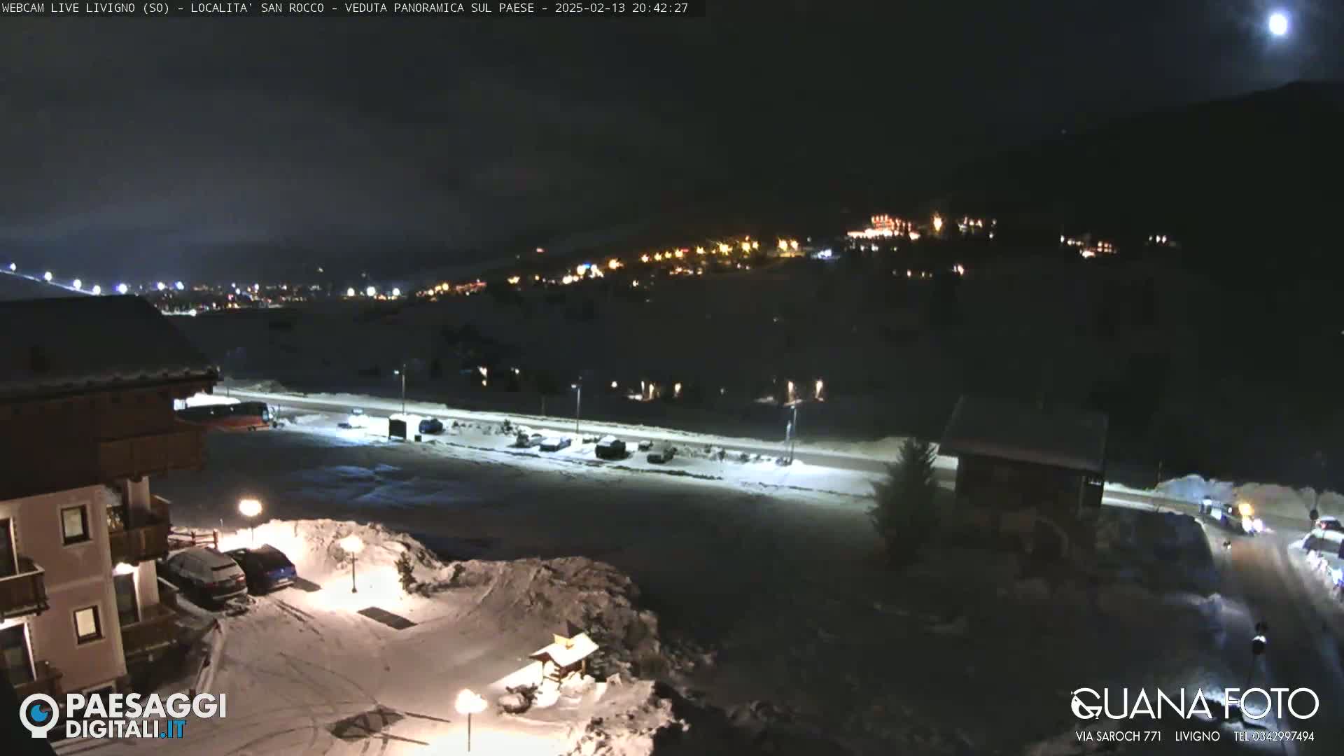 A nighttime, snow-covered landscape is viewed from an elevated position, showing a town's lights in the distance under a bright moon.