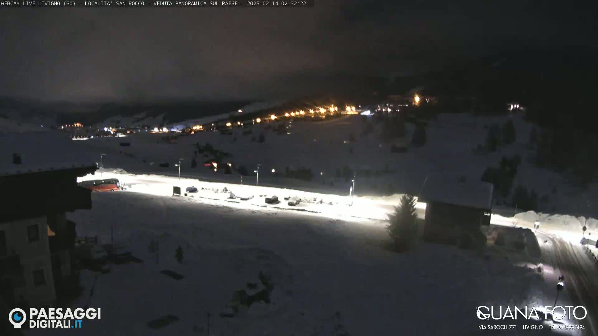 A snow-covered landscape at night, illuminated by artificial lights, shows a town in the distance and a few vehicles parked in a lot closer to the viewer.