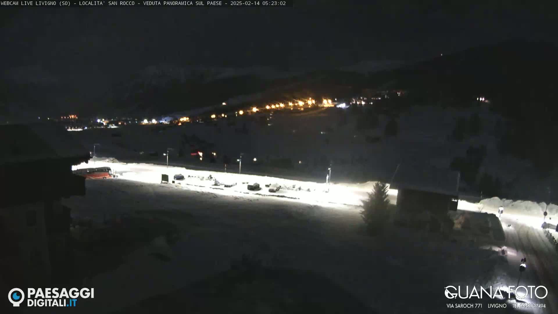 A nighttime, snow-covered scene shows a brightly lit area with parked vehicles and a bus, extending from a dark building toward a village with lights in the distance.