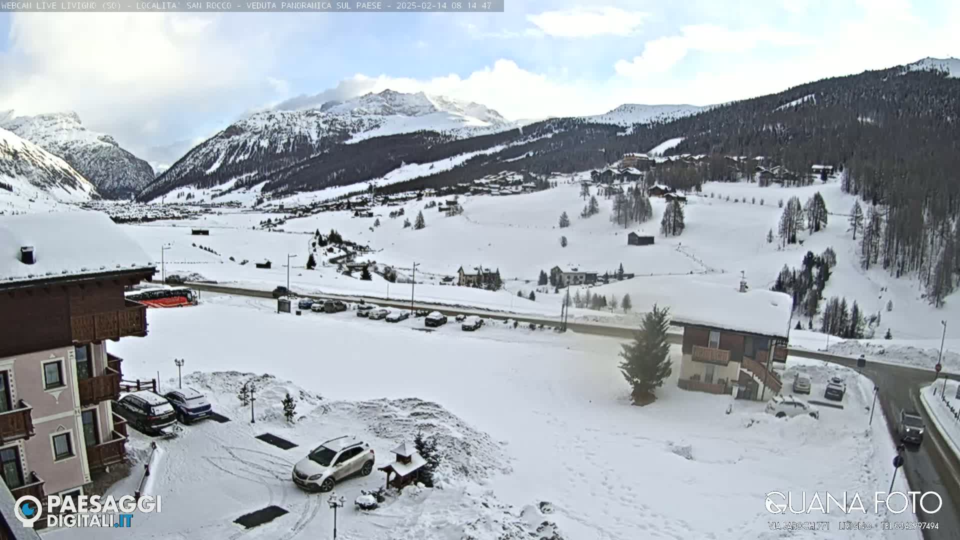 A snow-covered mountain village is viewed from a slightly elevated vantage point, under partly sunny skies.
