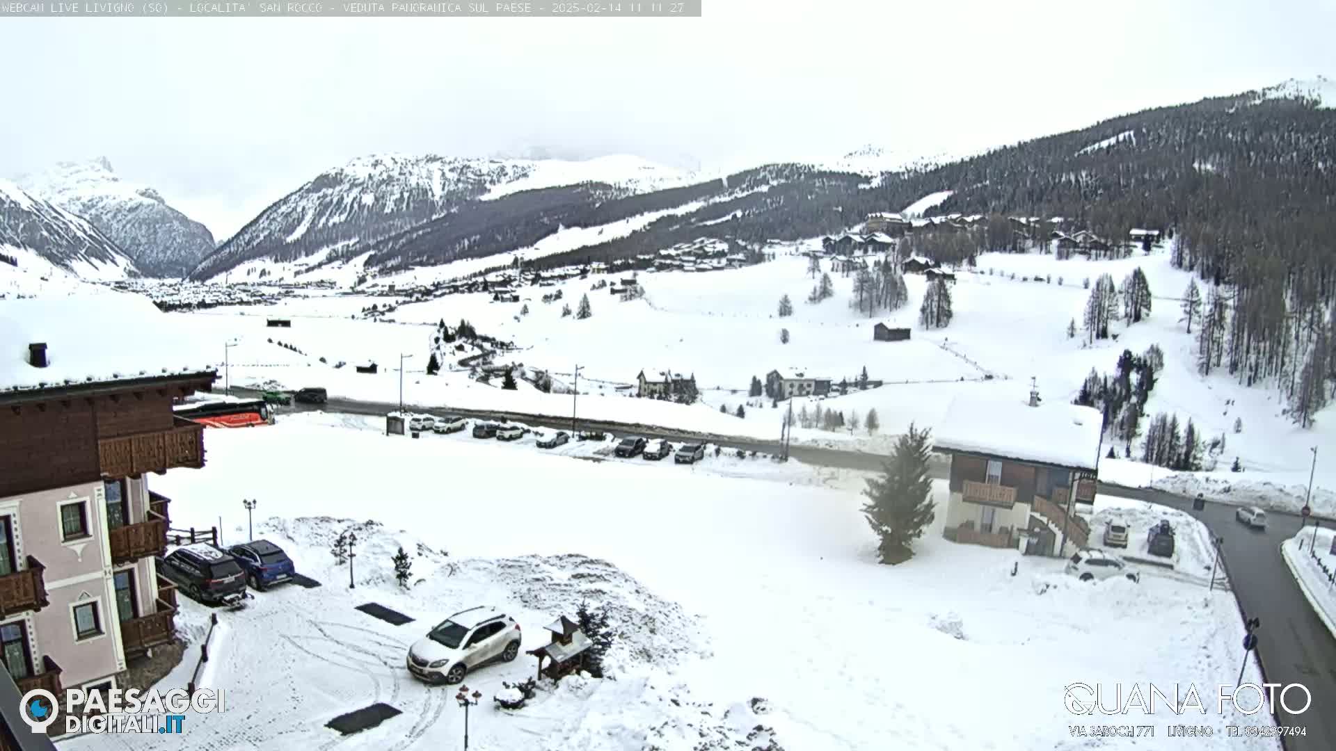 A snow-covered mountain village is viewed from a slightly elevated position, with several cars parked in a lot near a building, under an overcast sky.