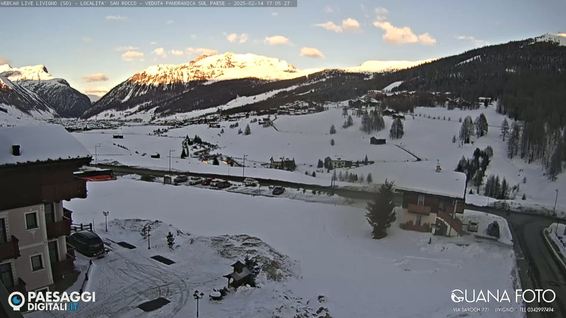 A snow-covered mountain landscape at sunset, with a village nestled in the valley below and buildings visible in the foreground.