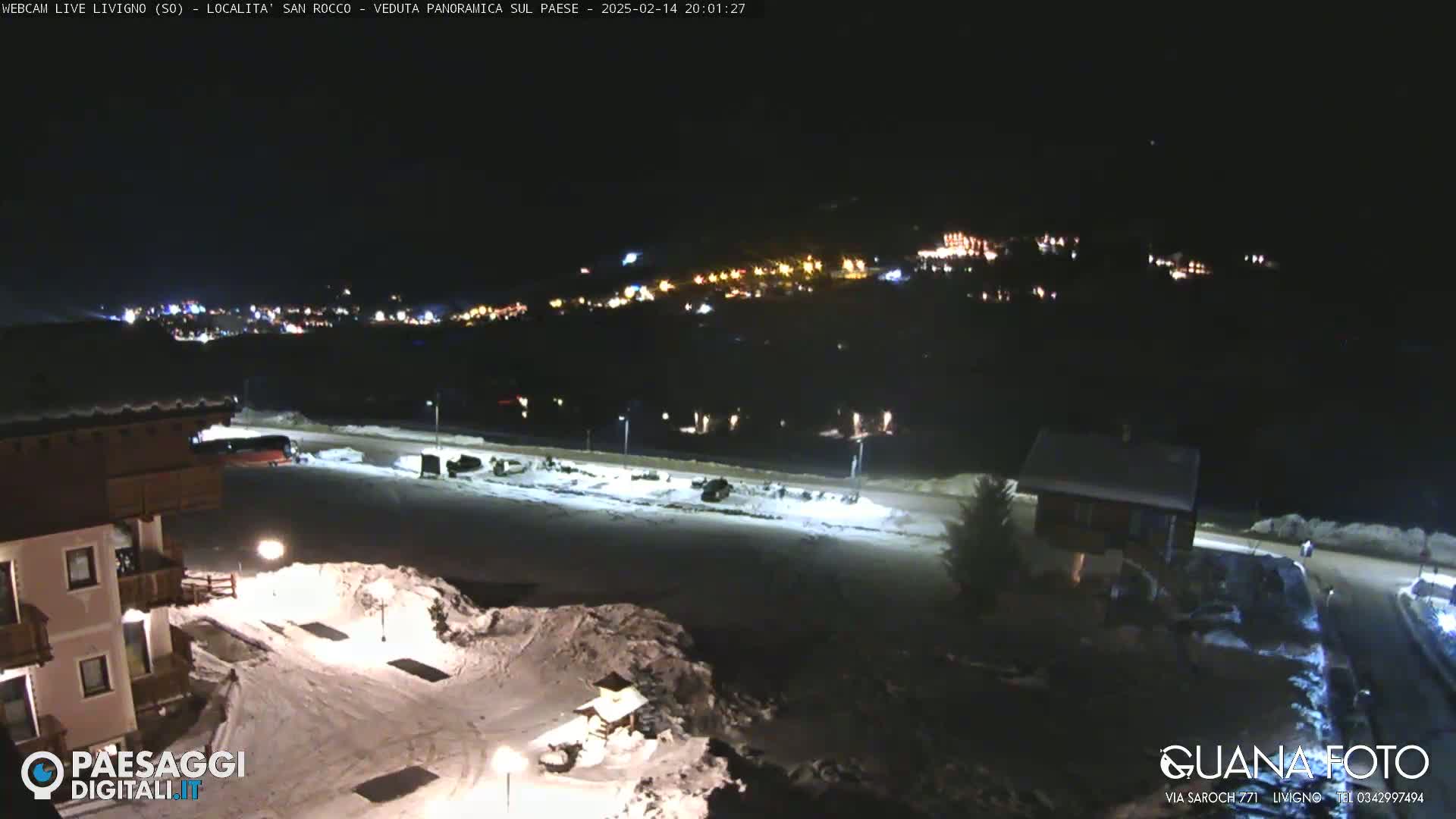 A nighttime, snow-covered scene shows a village in the distance illuminated by lights, with a building and parking lot in the foreground under a clear, dark sky.