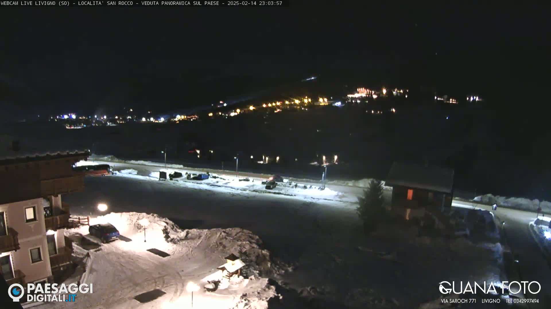 A nighttime, snow-covered parking lot and village are illuminated by streetlights and building lights, with a hill in the background.
