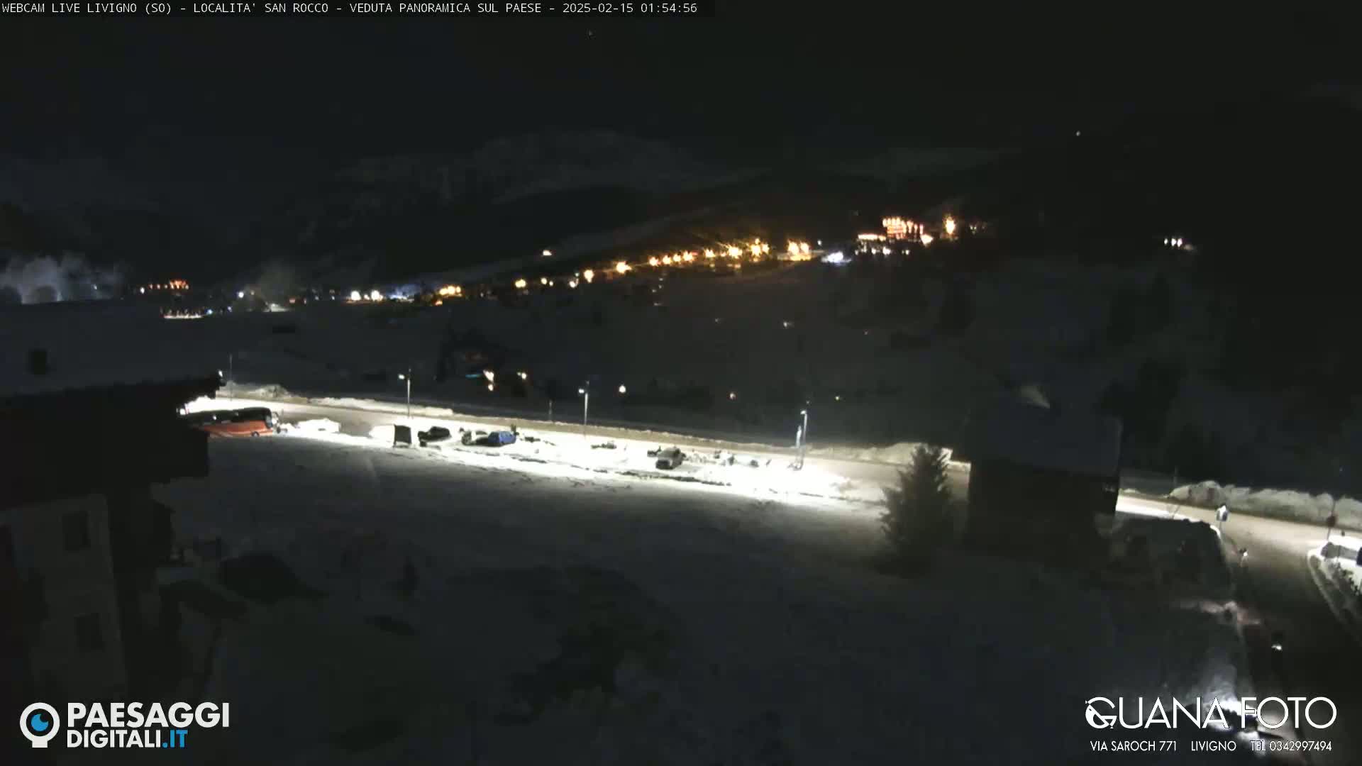 A nighttime, snow-covered scene shows a road with parked cars and a bus, leading to a village with illuminated buildings in the distance.
