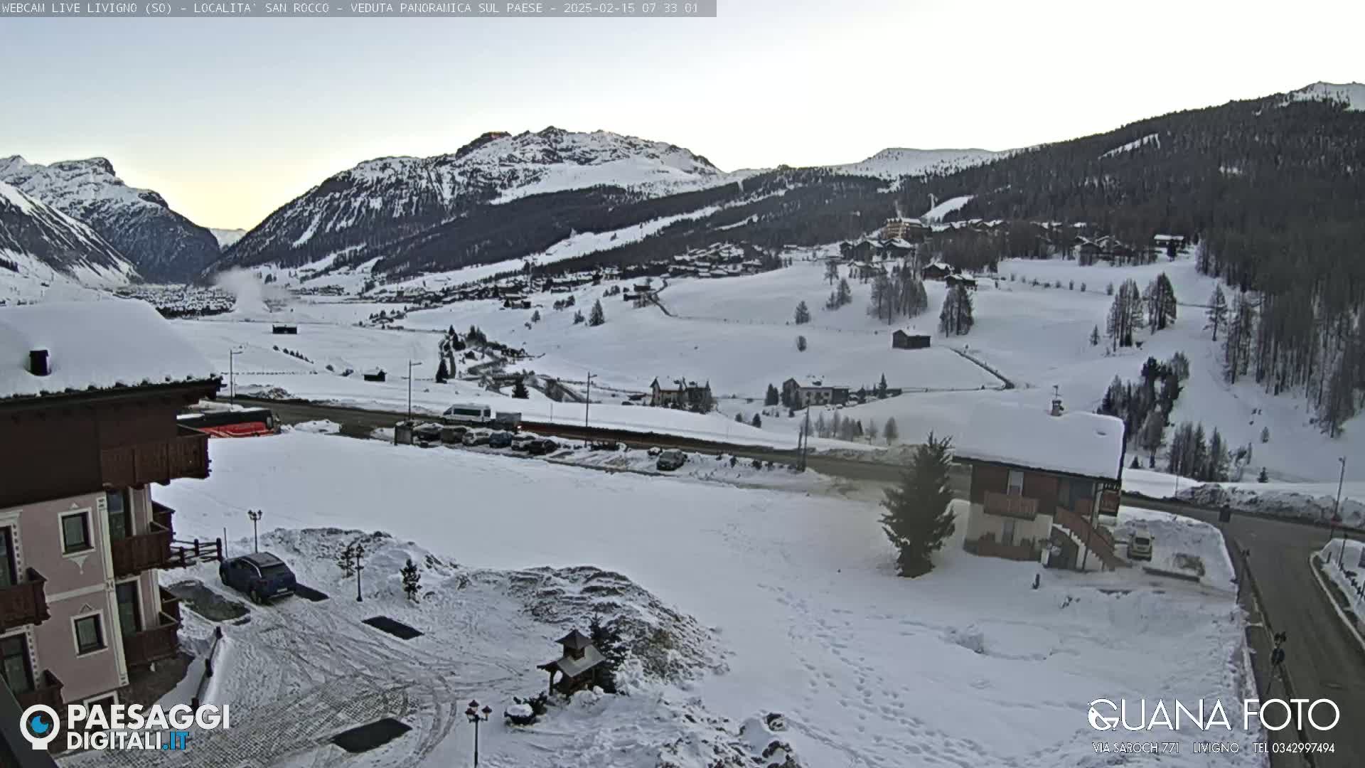 A snow-covered valley with buildings and mountains in the background under a clear, pale sky.