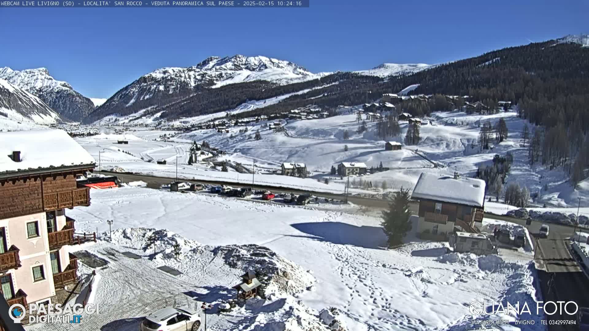 A snow-covered mountain valley scene on a sunny day, with buildings and vehicles visible, shows a picturesque winter landscape.