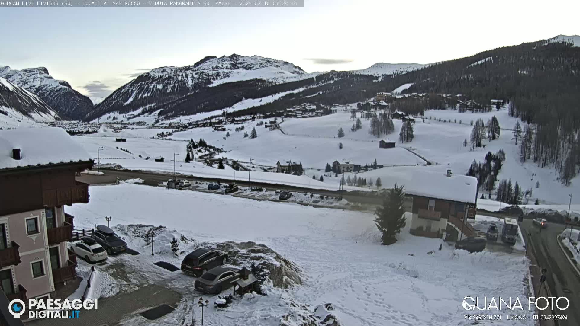 A snow-covered mountain valley village is viewed from a hillside, with several cars parked near a building in the foreground, under a twilight sky.