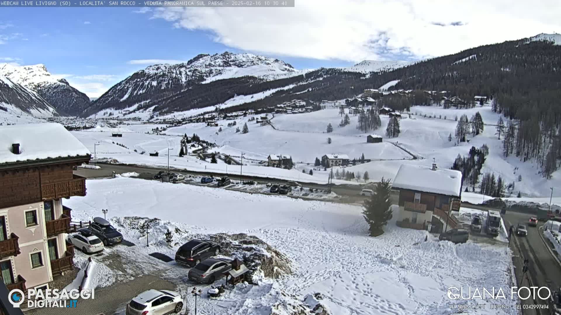 A snow-covered mountain valley town is seen from a high vantage point on a partly cloudy day; several cars are parked in a lot in the foreground.
