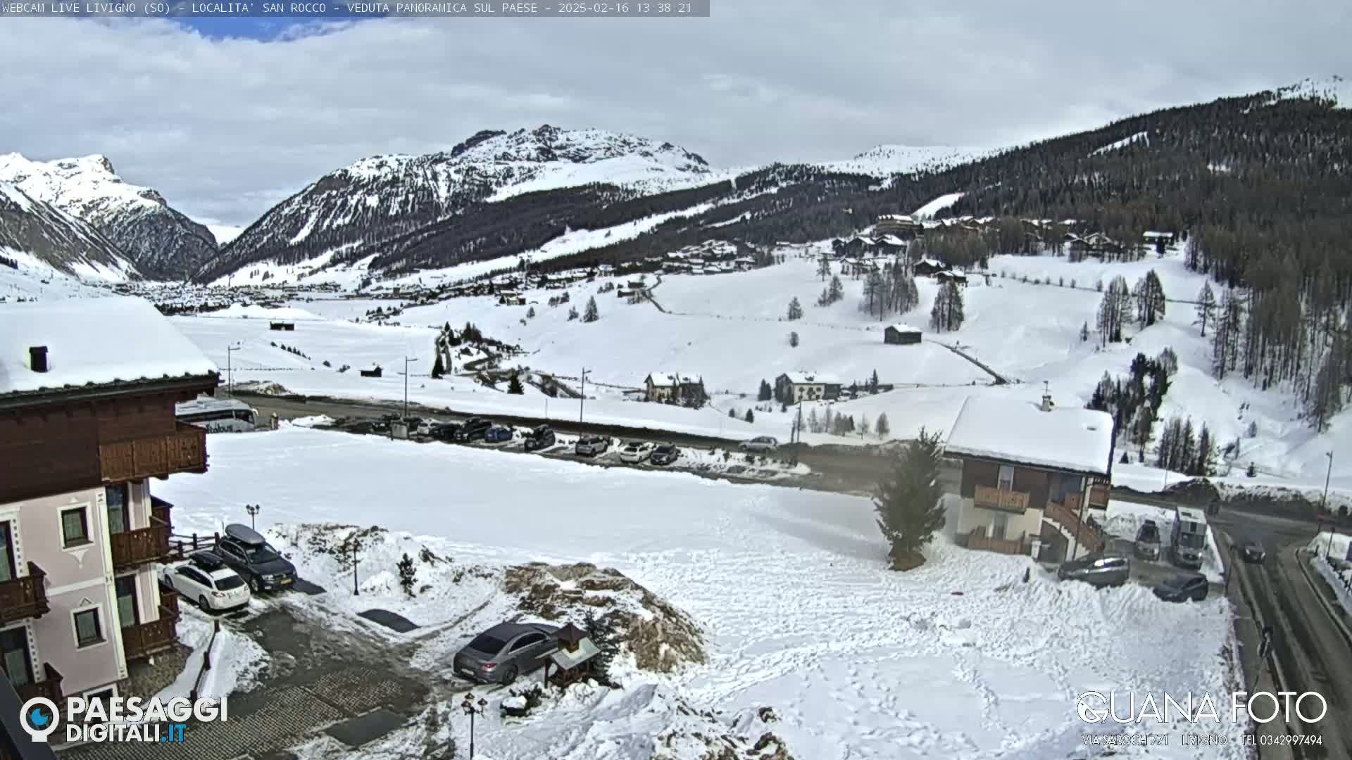 A snow-covered mountain village is viewed from a slightly elevated position on an overcast day, with cars parked in a lot near a building in the foreground.