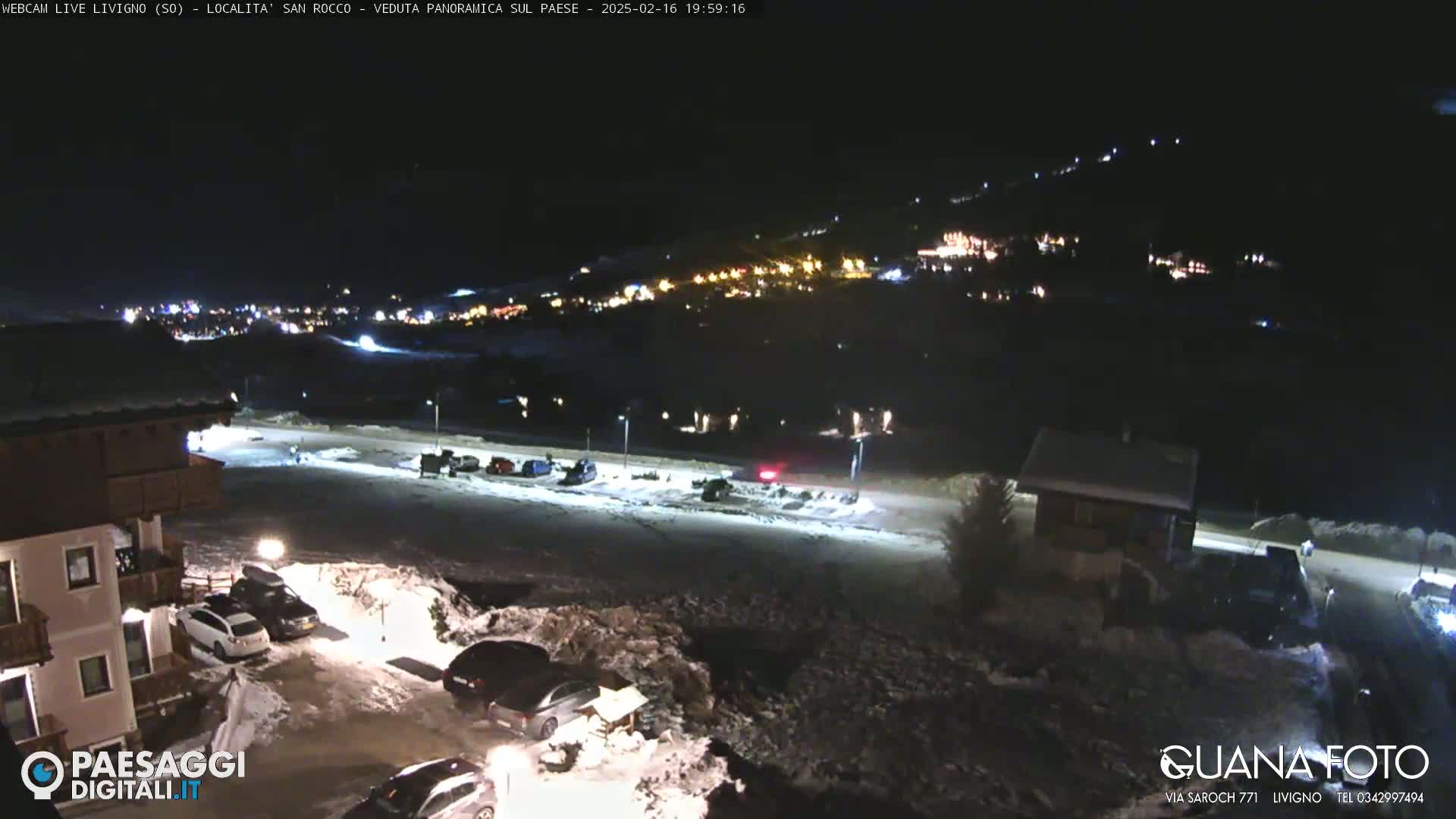 A nighttime, snow-covered parking lot and a small village illuminated by lights are seen in the distance.