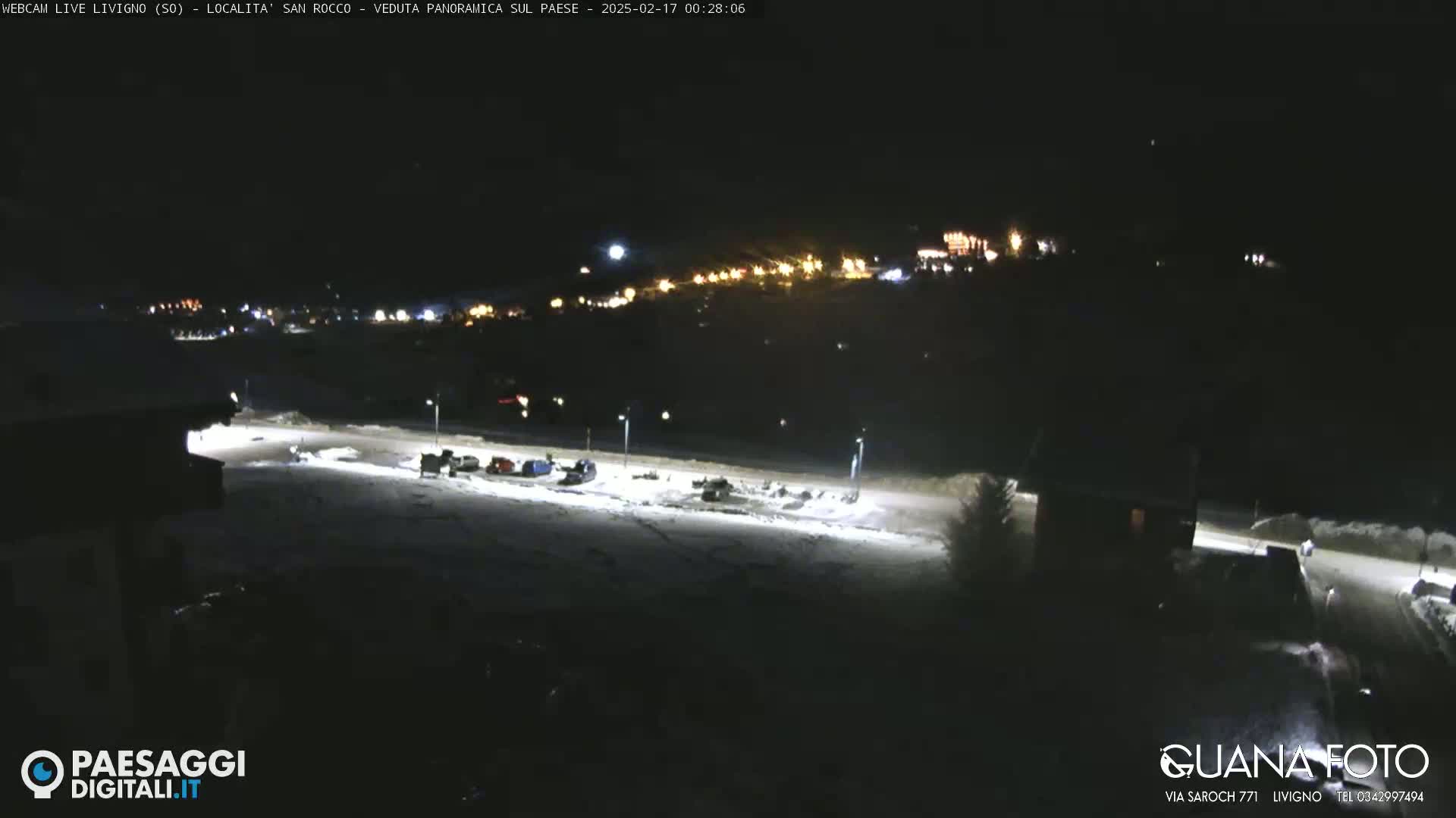 A nighttime, snow-covered parking lot with a few vehicles is illuminated by artificial lights, overlooking a dimly lit town nestled on a hillside.