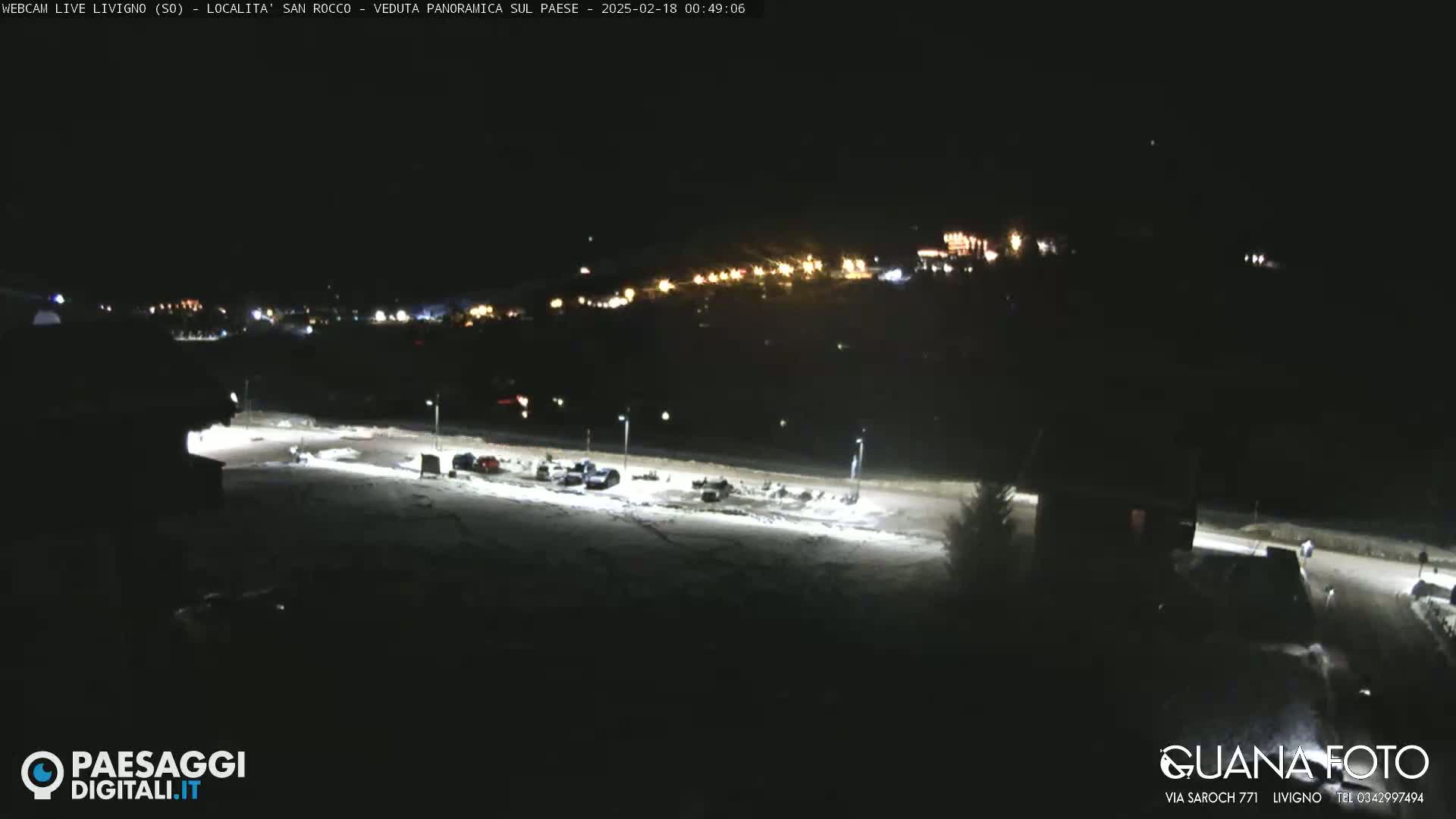 A nighttime, snow-covered parking lot with a few vehicles is visible, and a small town with lights is in the distance under a clear, dark sky.