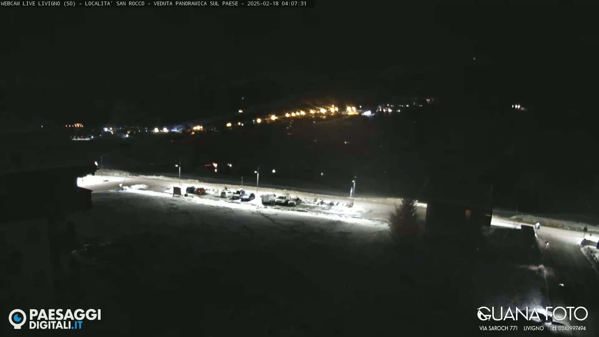 A nighttime, snow-covered parking lot with a few vehicles and distant lights from a town in the background.