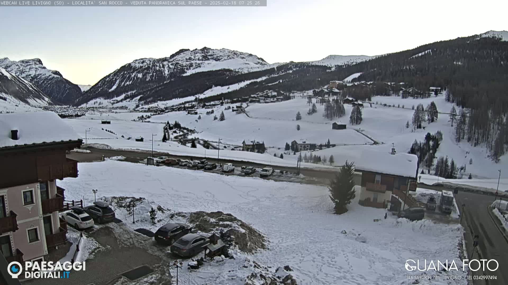 A snow-covered mountain valley with buildings and parked cars, under a clear, light sky.