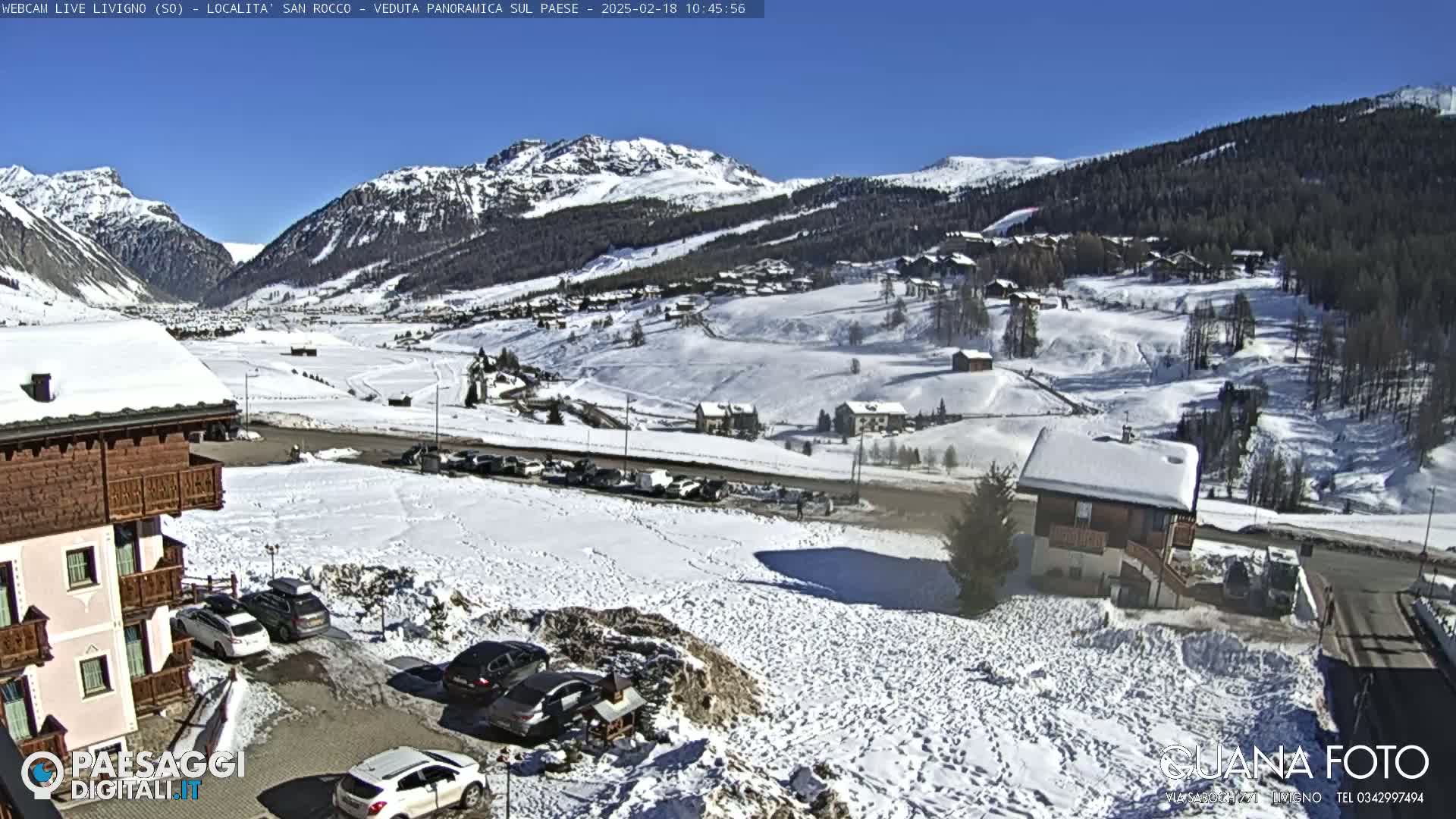 A snow-covered mountain village is viewed from a hillside on a sunny day, with cars parked in a lot near buildings.